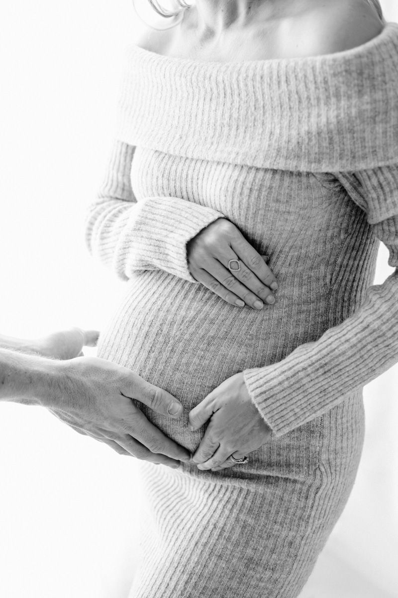 Black and white image showing hands gently cradling a pregnant belly in a cosy ribbed sweater dress.