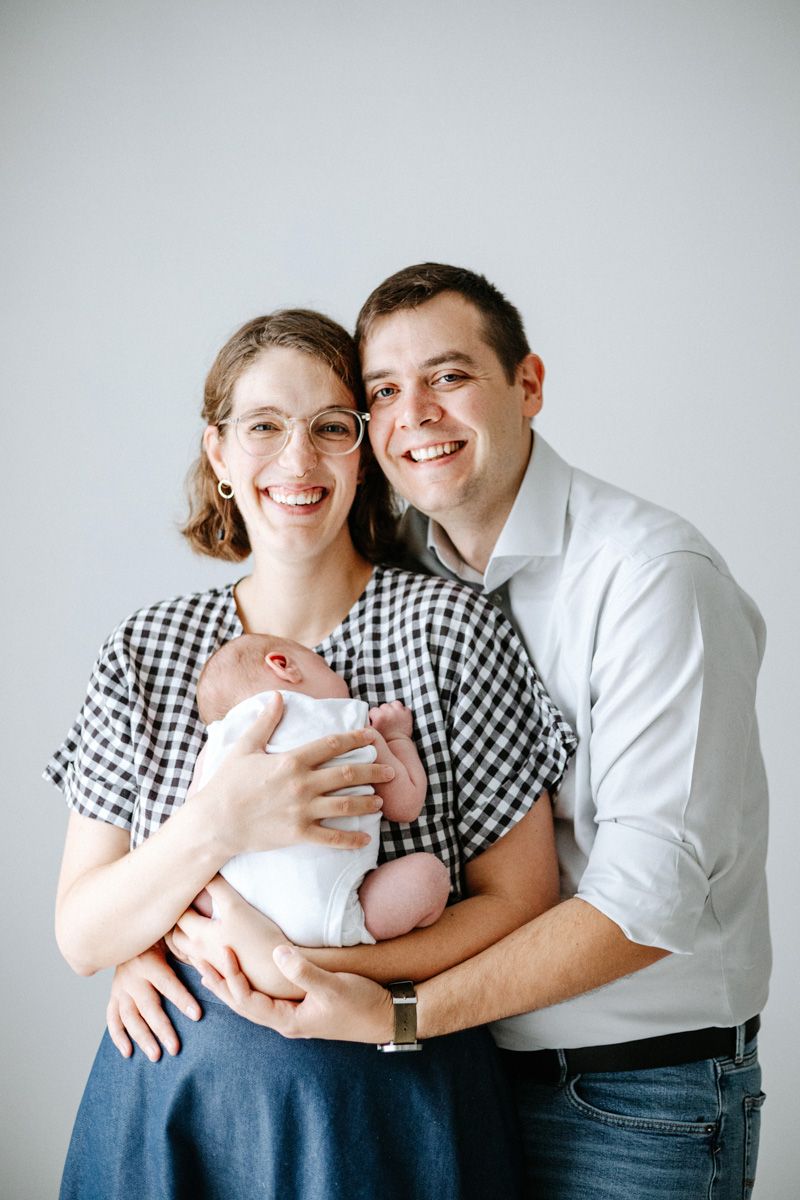 A couple in gingham and white dress shirts pose lovingly with their newborn baby against a light gray backdrop.