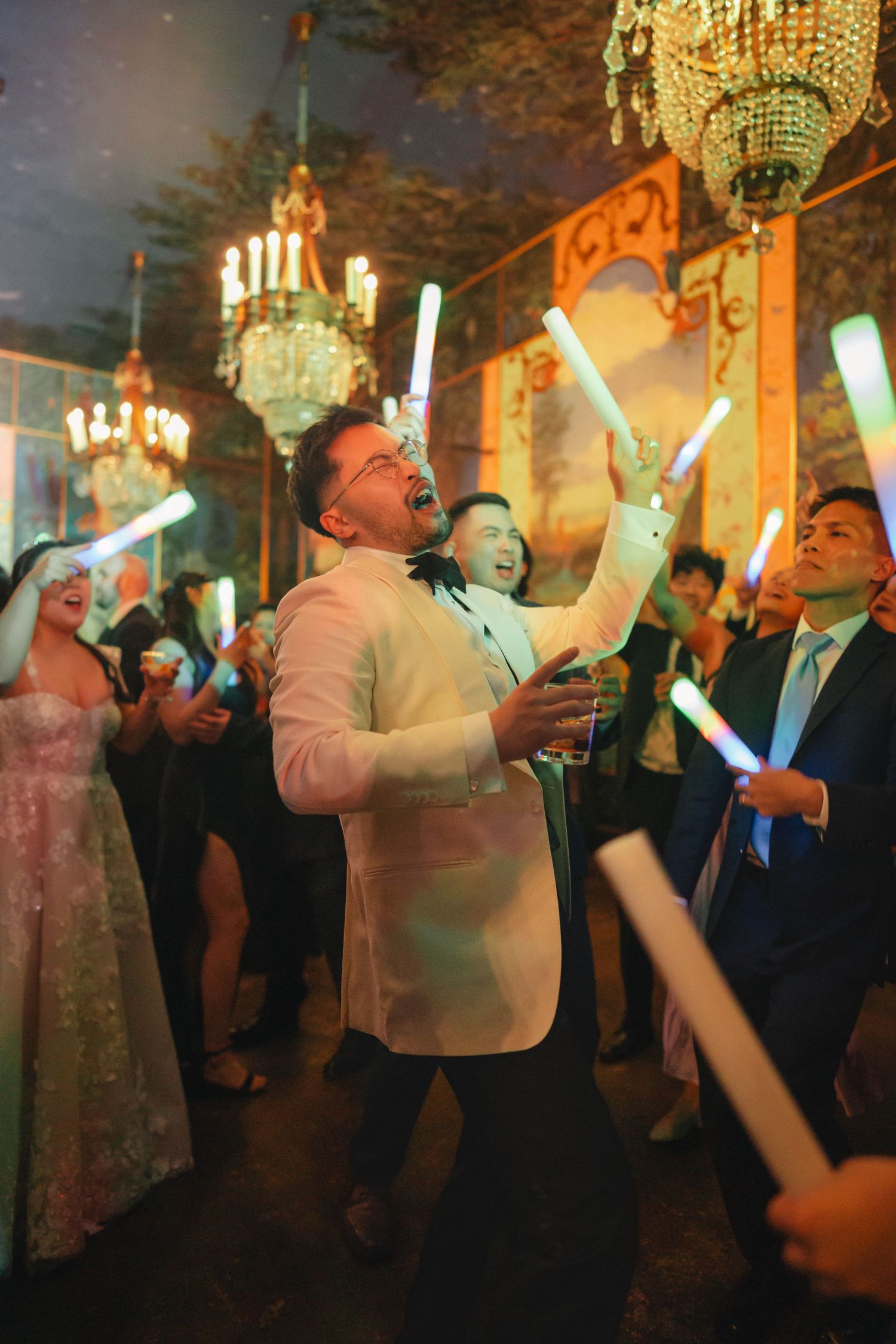 Groom in white tuxedo dancing joyfully with glowing light sticks at elegant wedding reception with chandeliers.