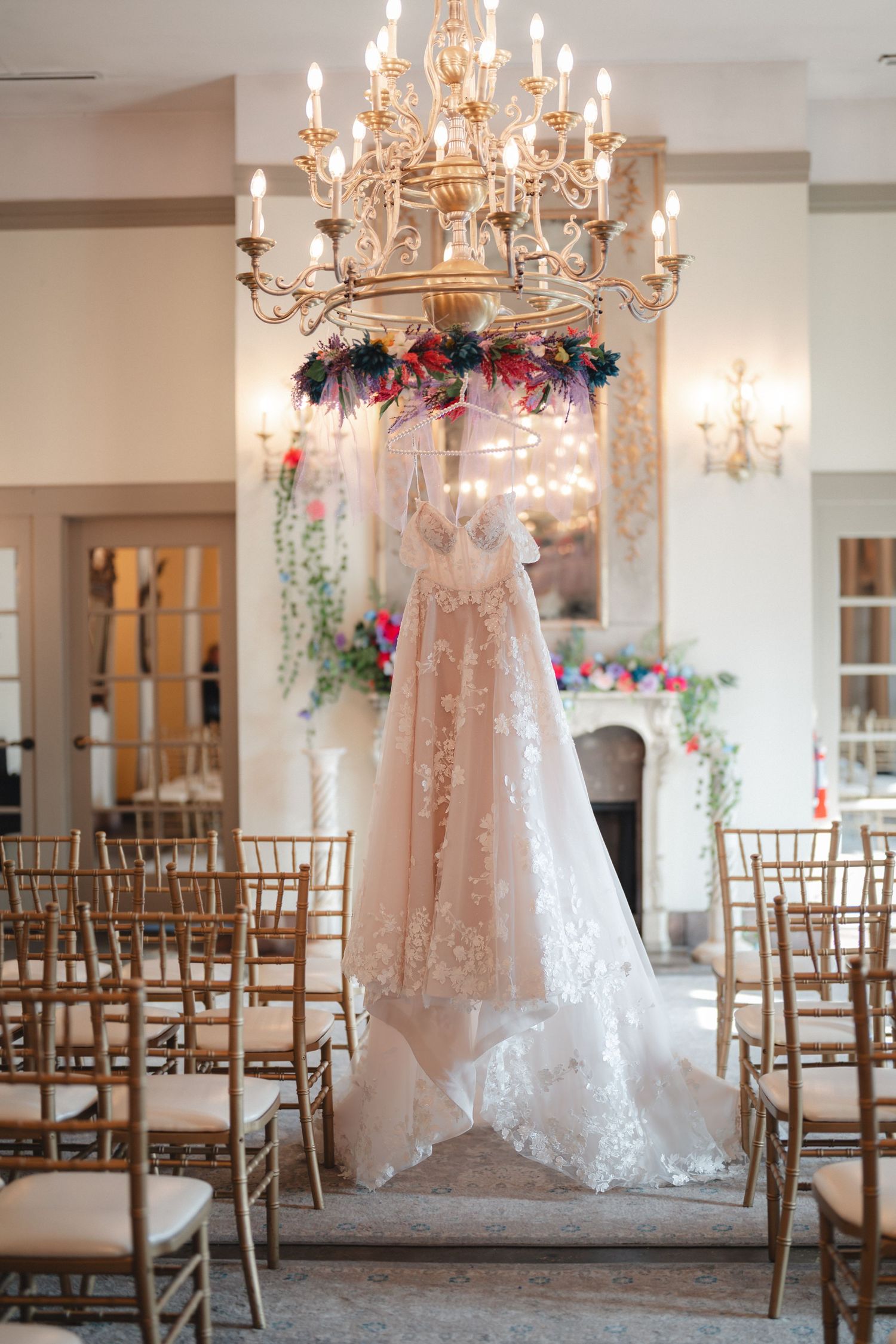 Elegant wedding dress hanging from a gold chandelier adorned with flowers in a romantic venue with gold chiavari chairs.