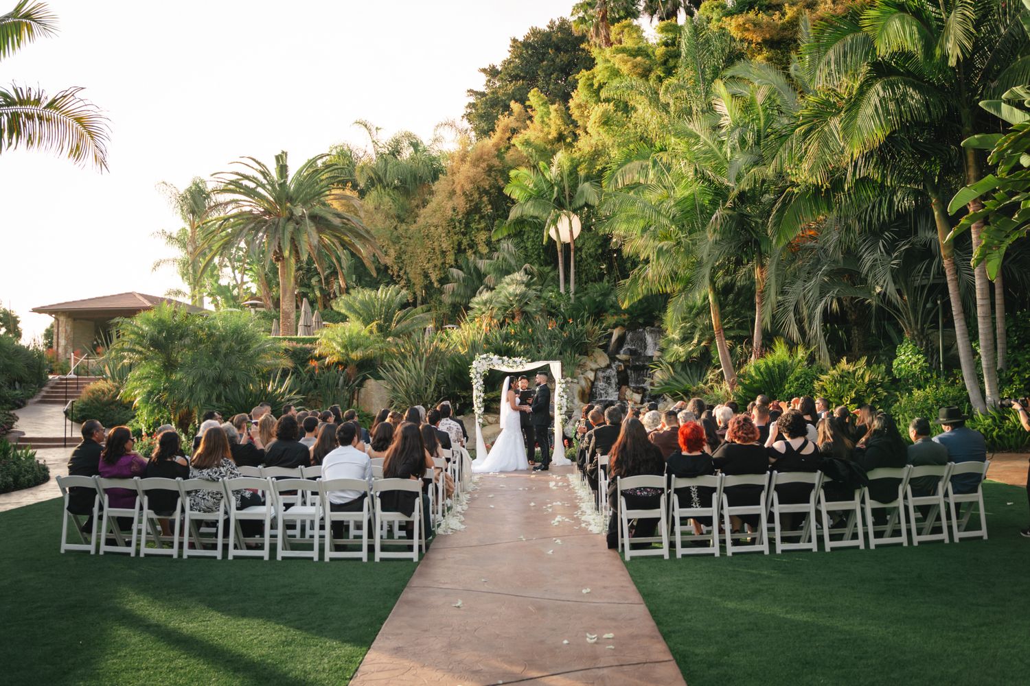 Outdoor wedding ceremony setup with white chairs arranged in rows along a palm tree-lined path at sunset.