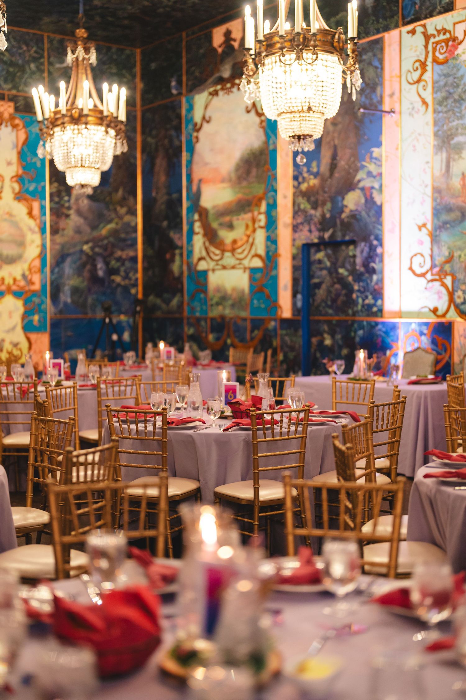 Elegant banquet hall with crystal chandeliers, gold chiavari chairs, white tables, red napkins, and colorful mural walls.