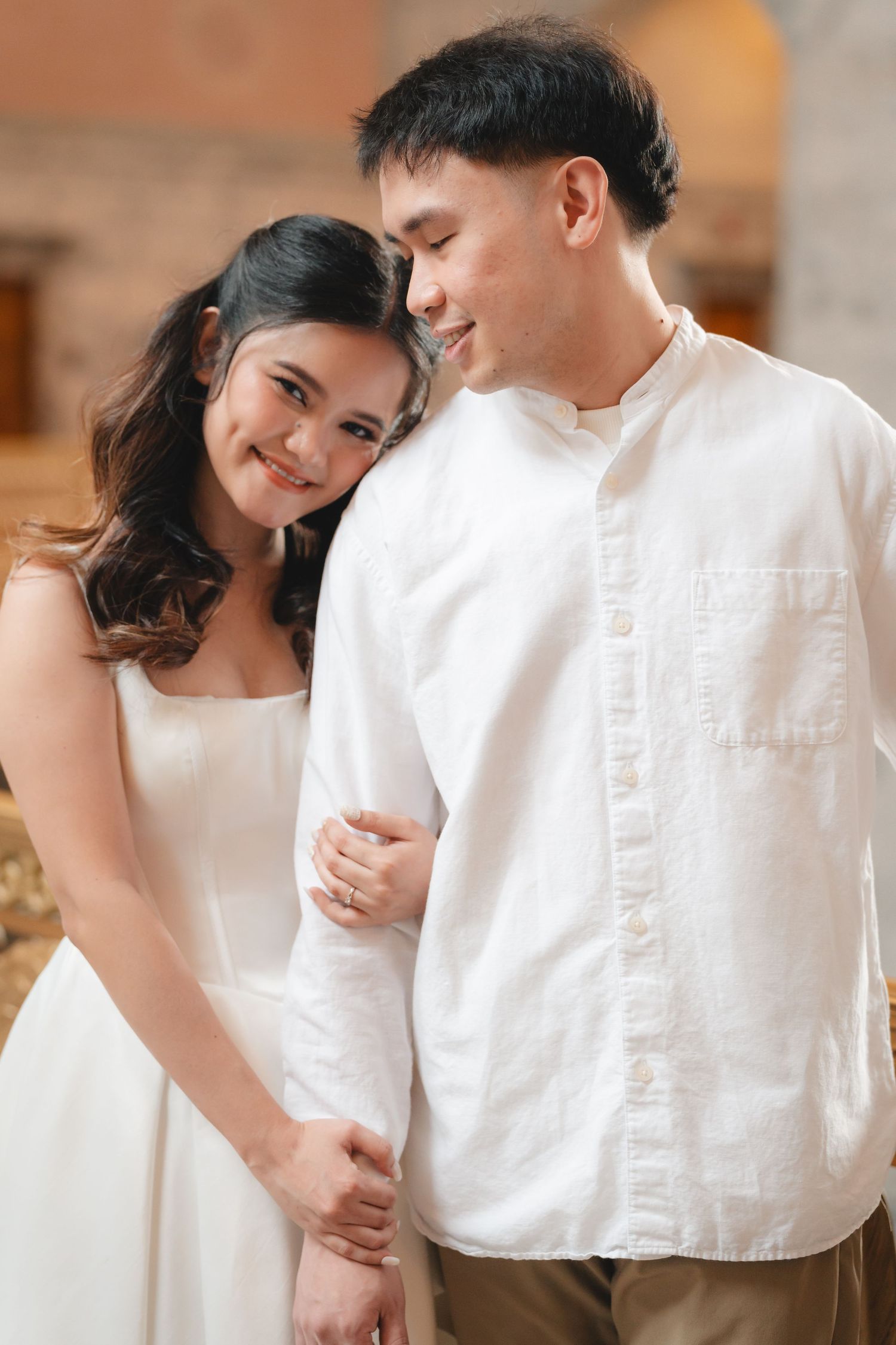 A couple in white clothing embraces intimately during a romantic photoshoot in a warmly lit indoor setting.