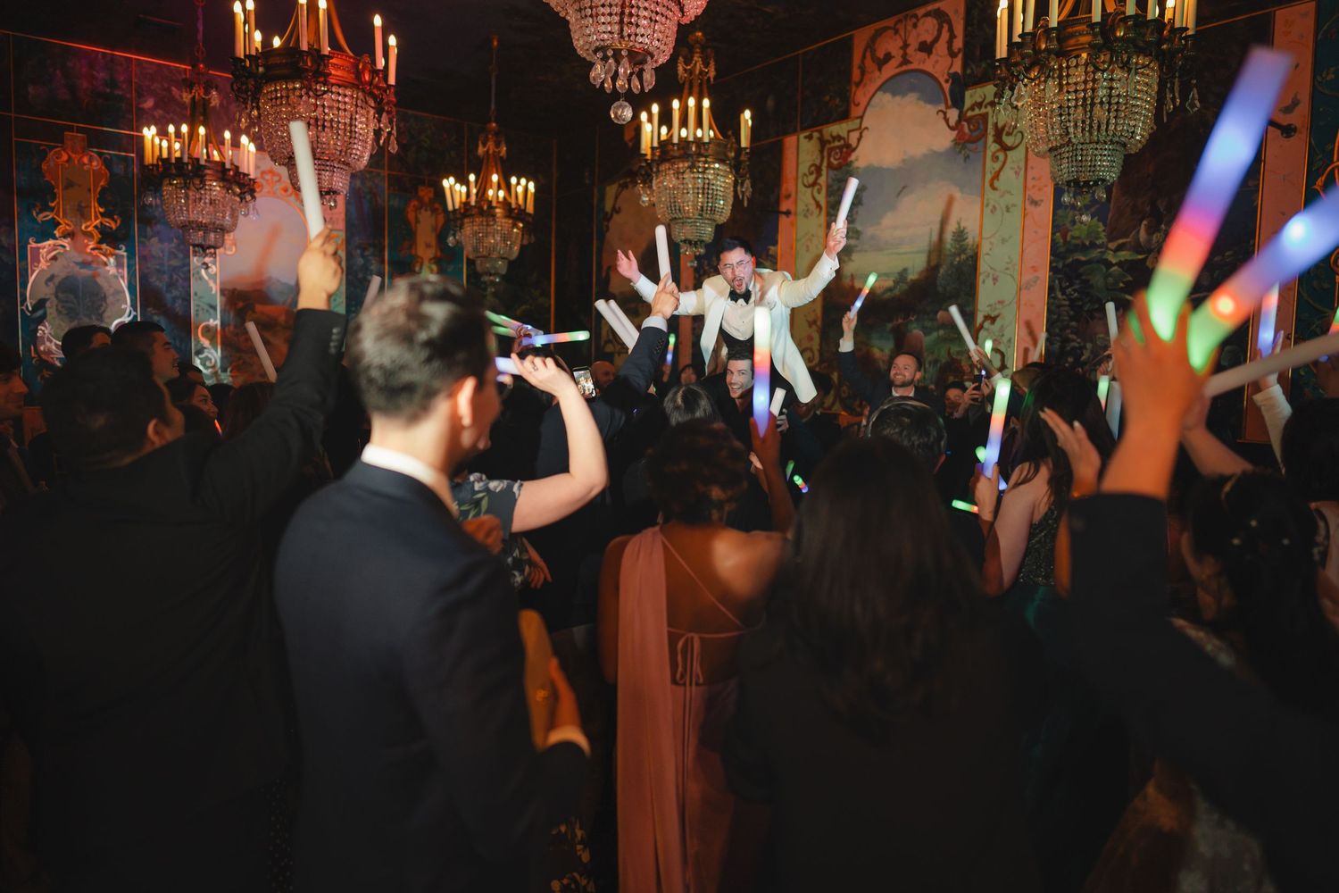 Guests celebrate at an elegant party with glowing light sticks in a ornate venue with chandeliers.