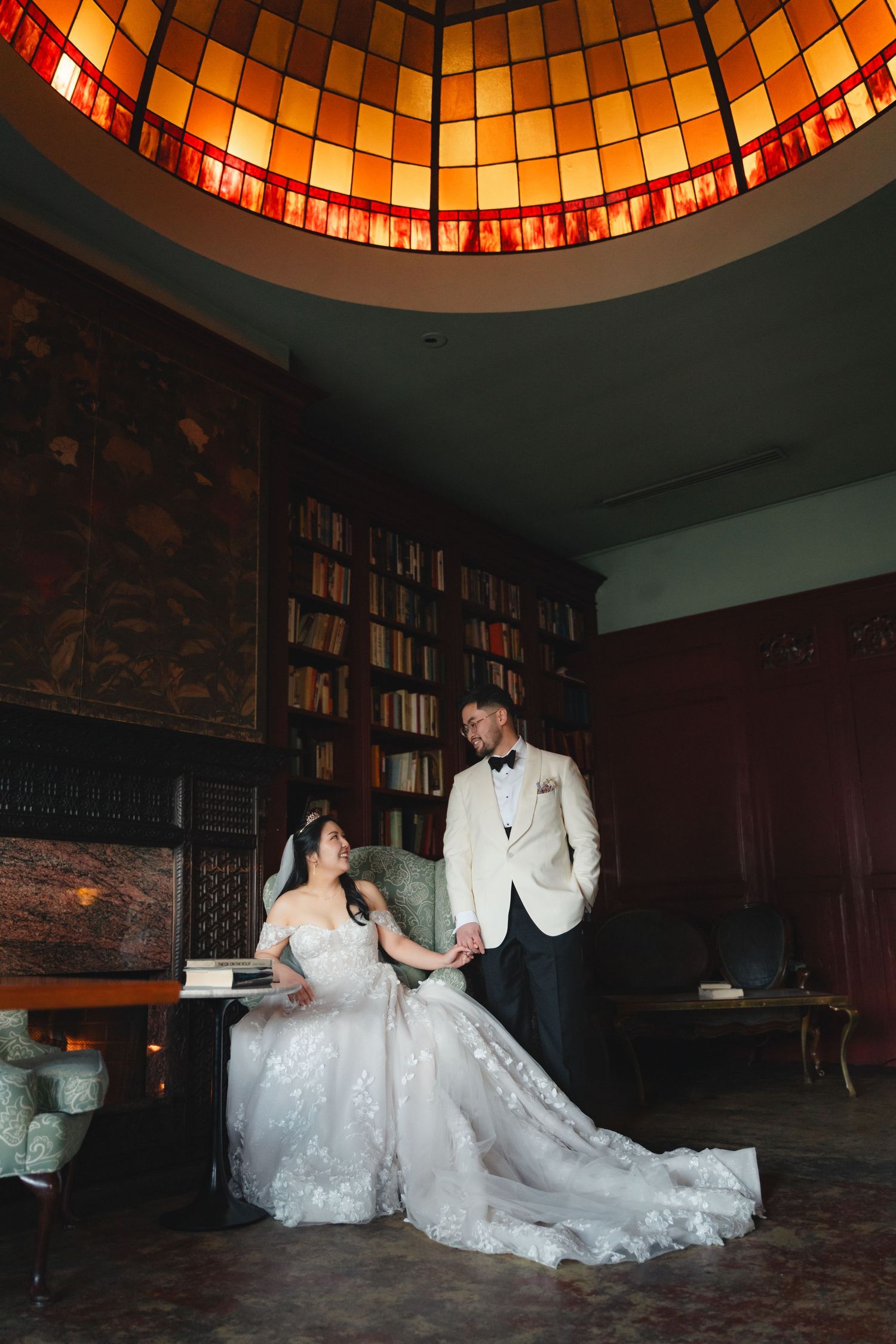 Bride in lace gown and groom in white tuxedo pose under stained glass dome in elegant library setting.