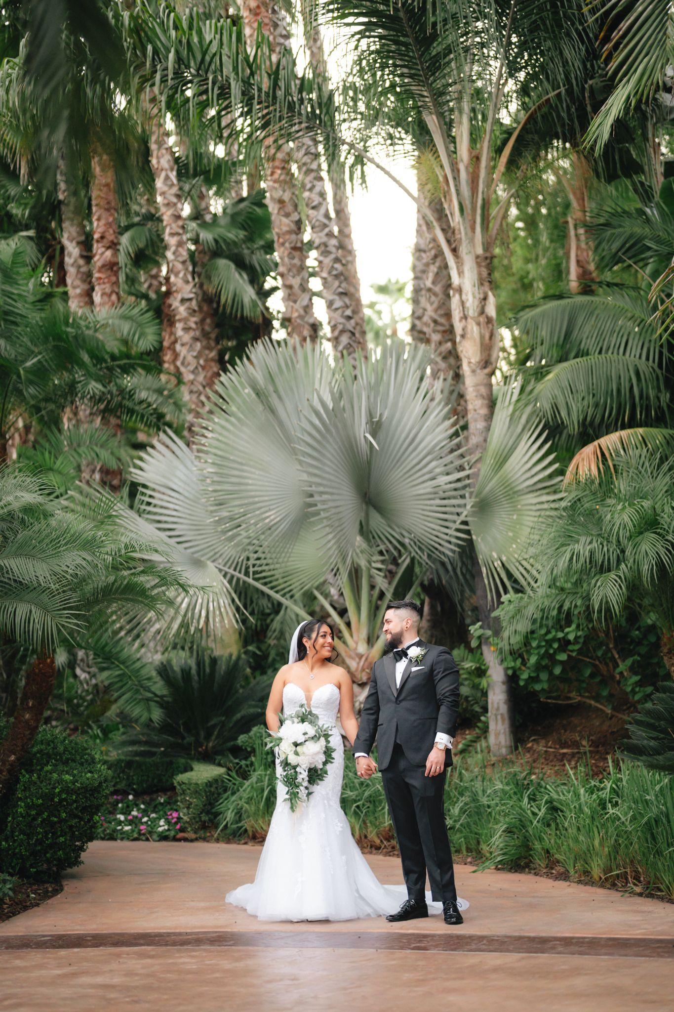 A romantic wedding couple poses in a lush tropical garden setting with palm trees and fan palms creating a natural backdrop.