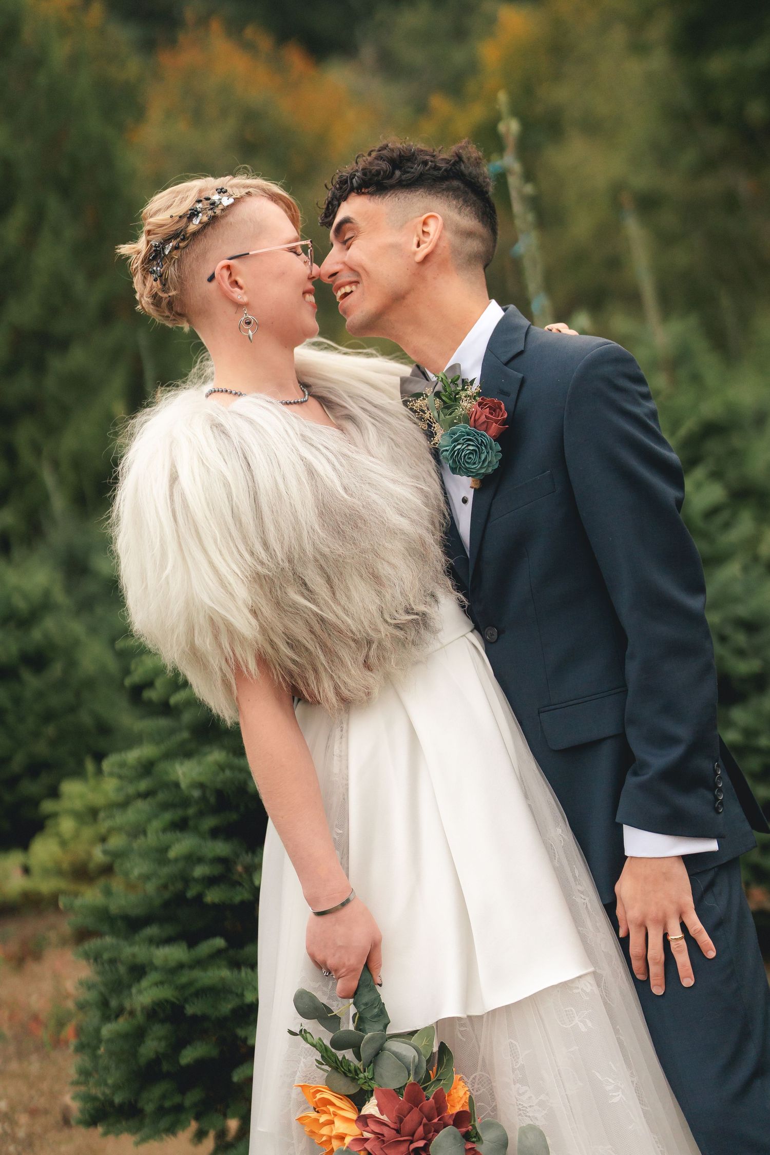 A couple in wedding attire share a romantic kiss in a garden setting with autumn foliage in the background.