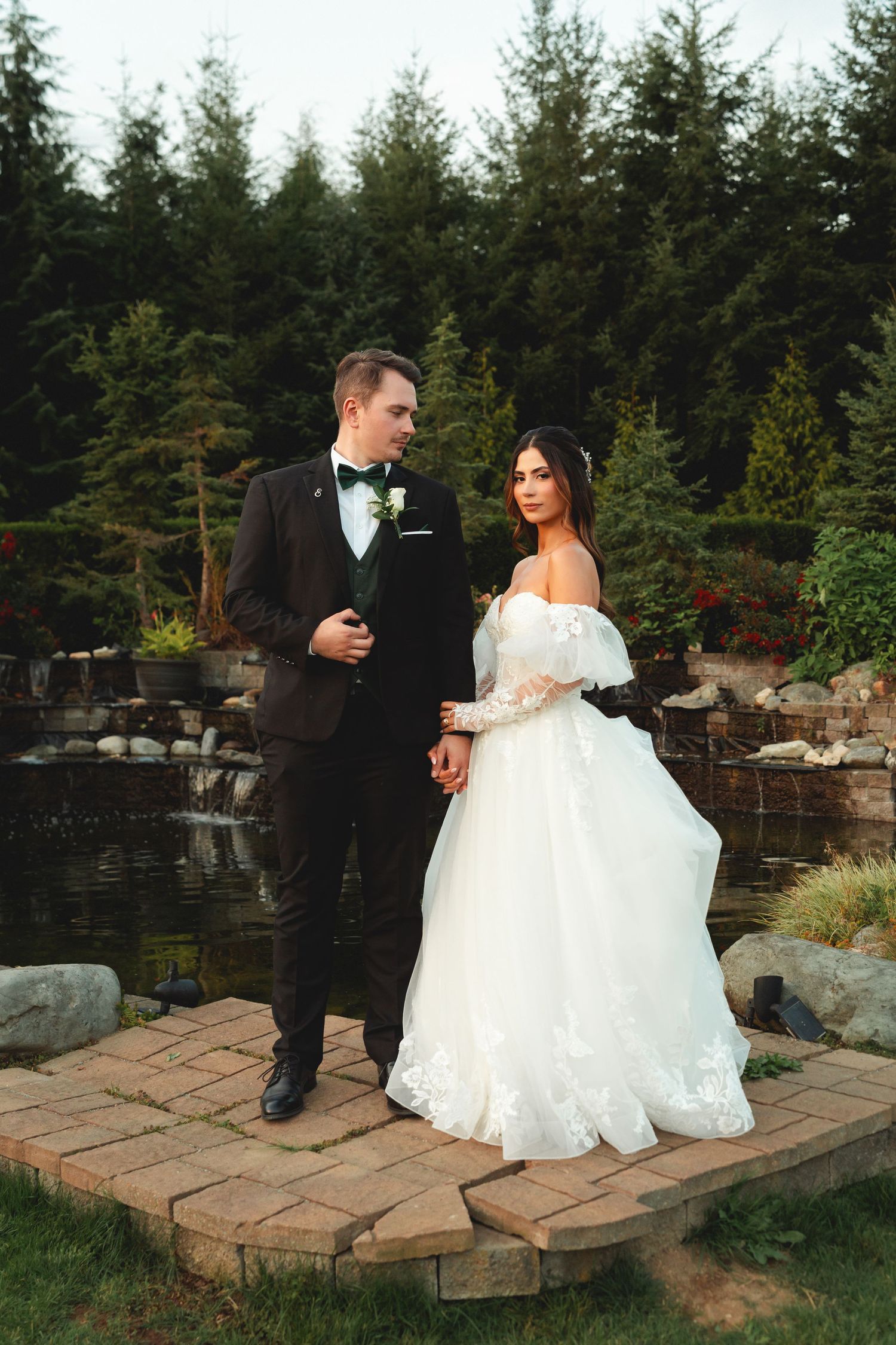 Couple in formal wedding attire sharing a romantic moment on stone steps surrounded by evergreen trees and landscaping.