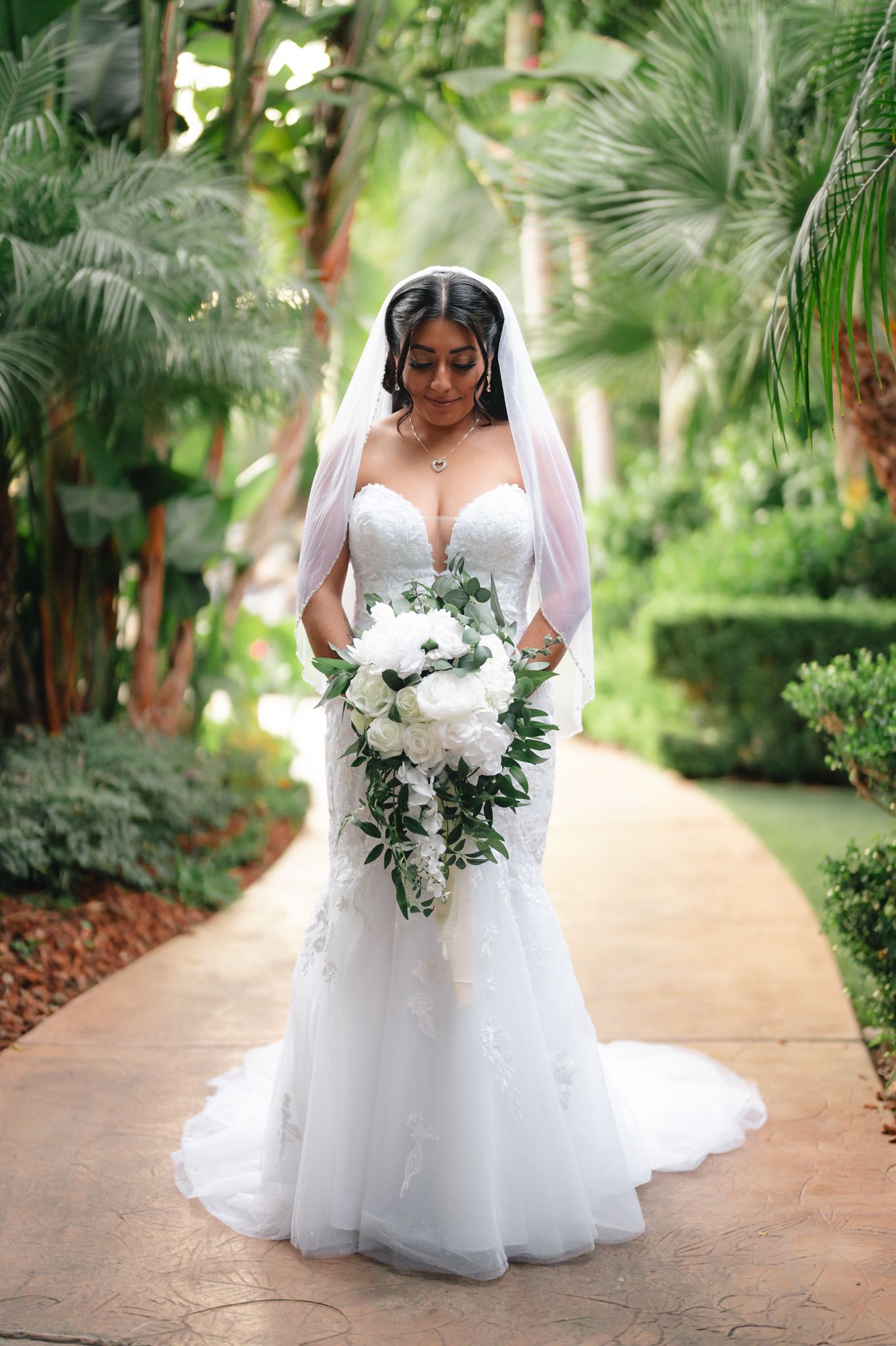 Beautiful white wedding gown with lace details and flowing veil photographed in a tropical garden setting.
