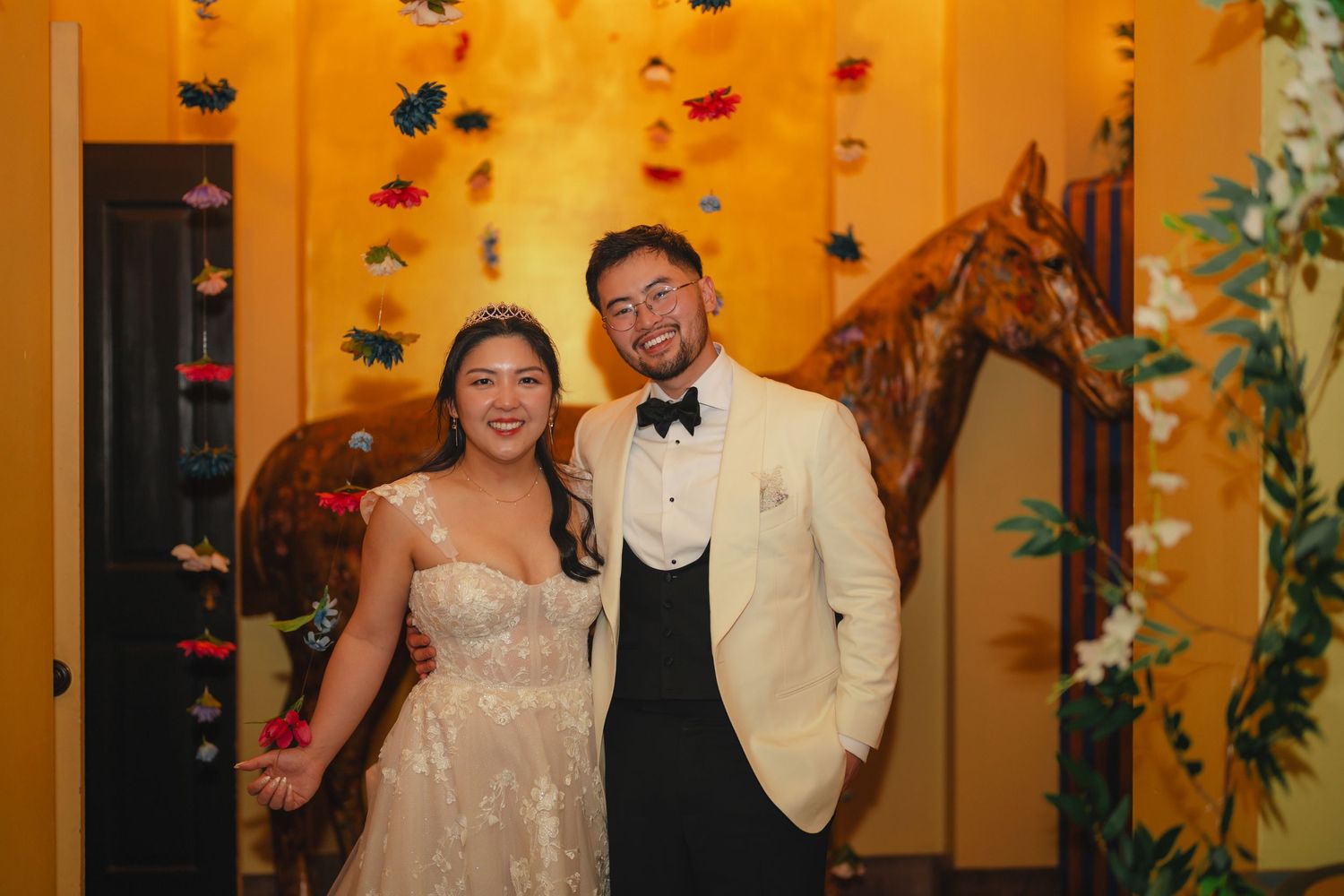 A smiling bride in a lace gown and groom in white tuxedo pose together at a floral-decorated wedding venue.