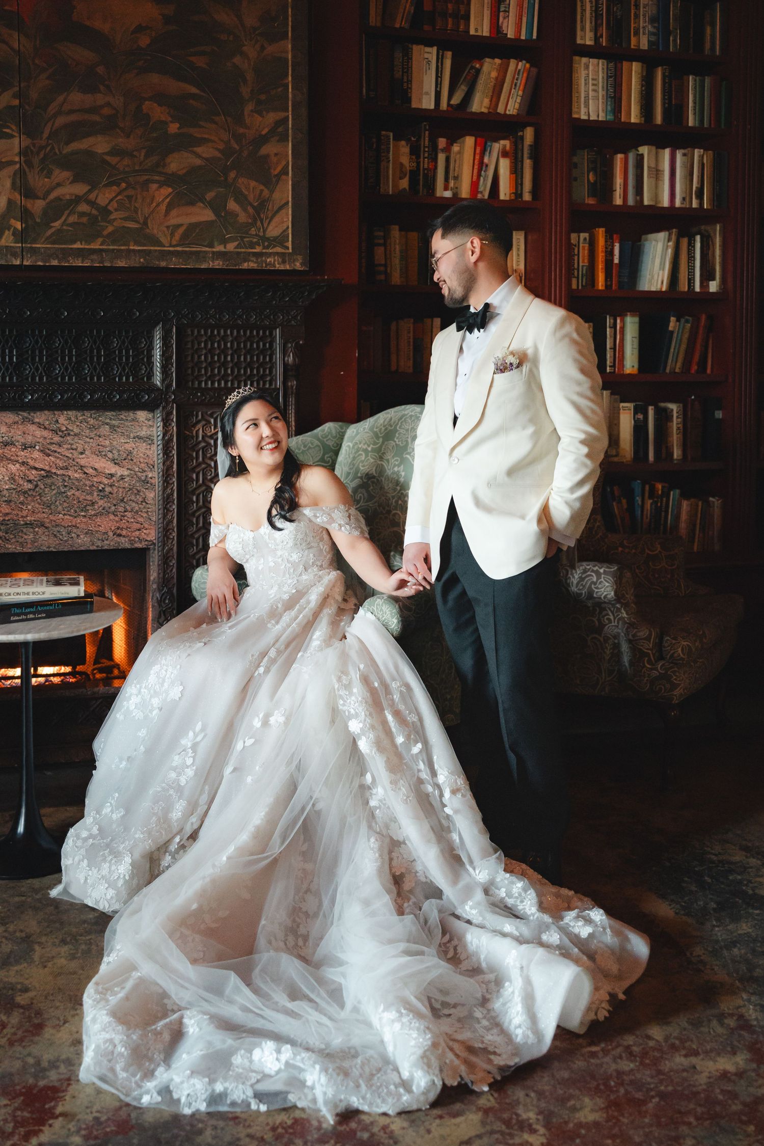 Bride in lace ballgown sits by fireplace as groom in white tuxedo jacket holds her hand in elegant library.