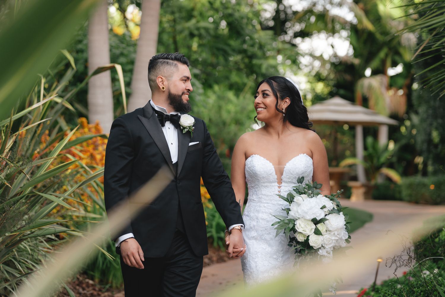 Couple in formal wedding attire walking along lush garden path with tropical foliage.