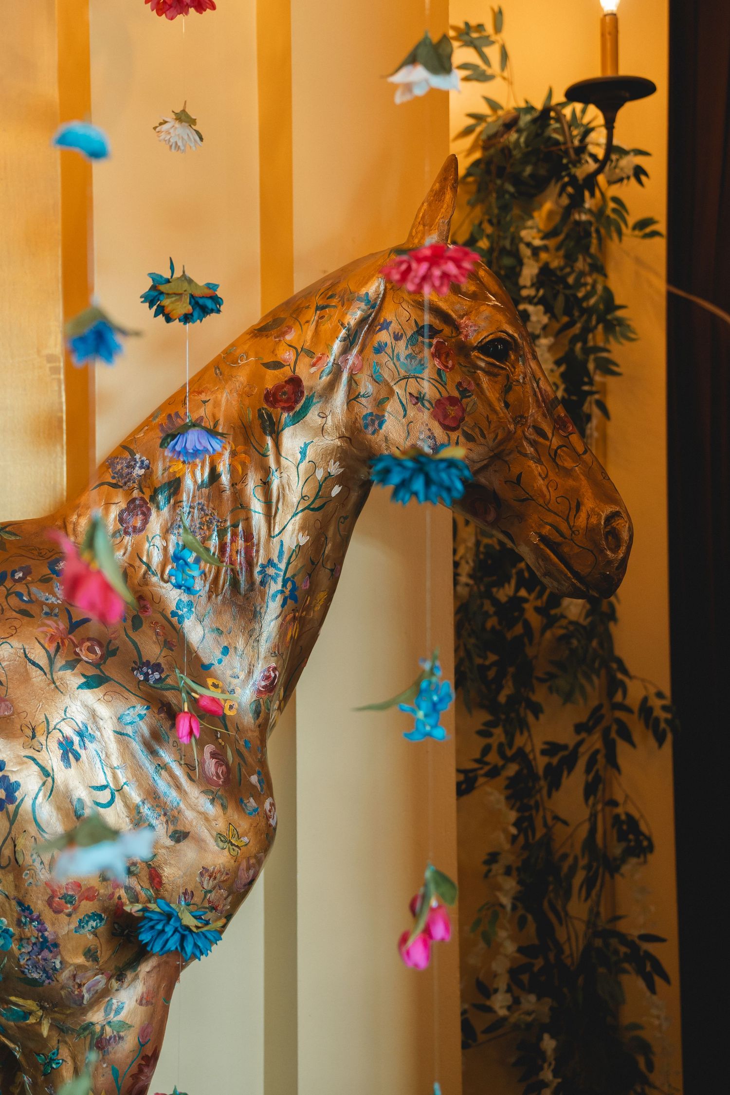 Decorative horse sculpture adorned with floral paintings and colorful hanging paper flowers against a warm yellow wall.