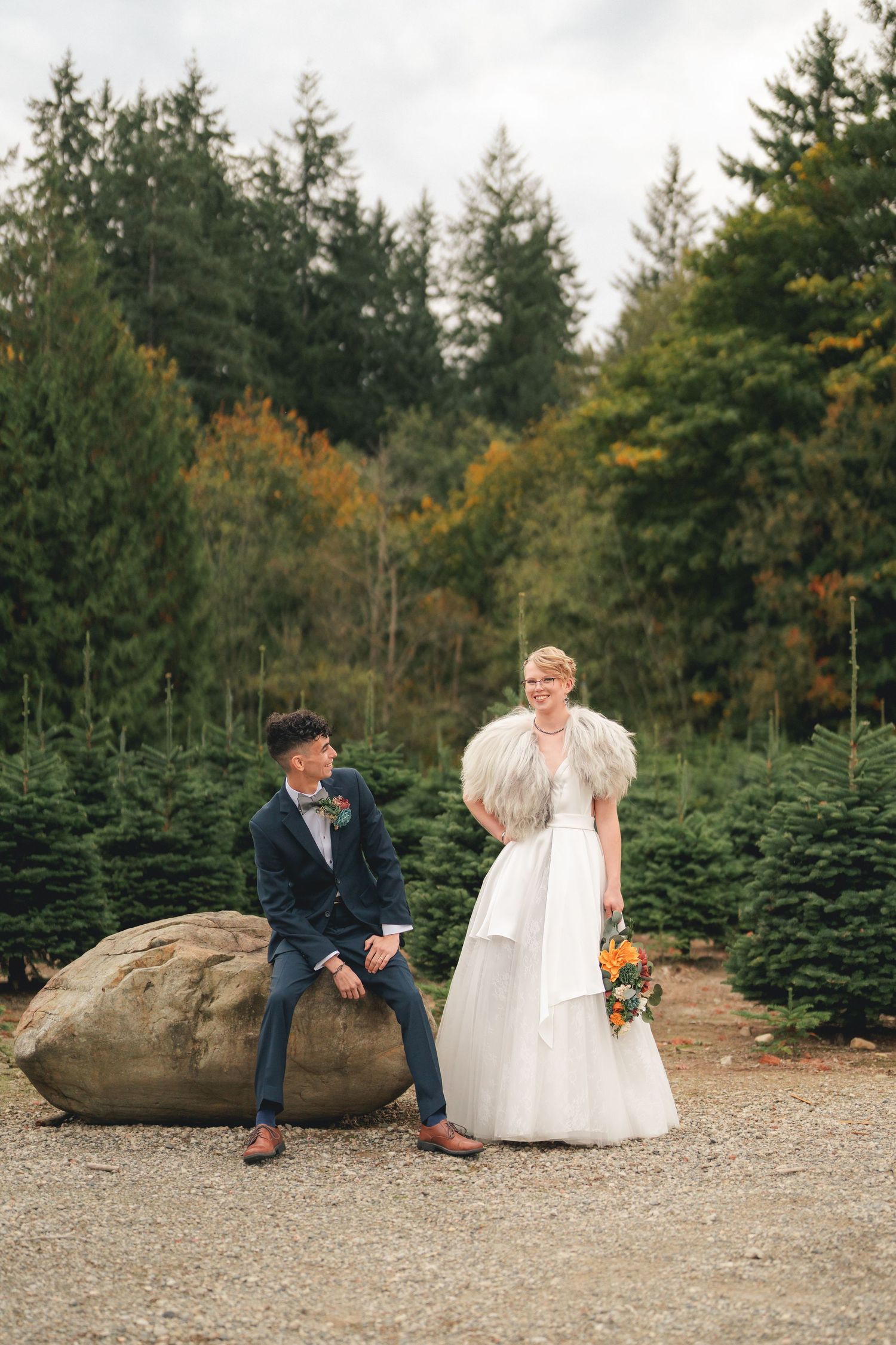 A couple sits together on a large rock at a Christmas tree farm during a fall photo session.