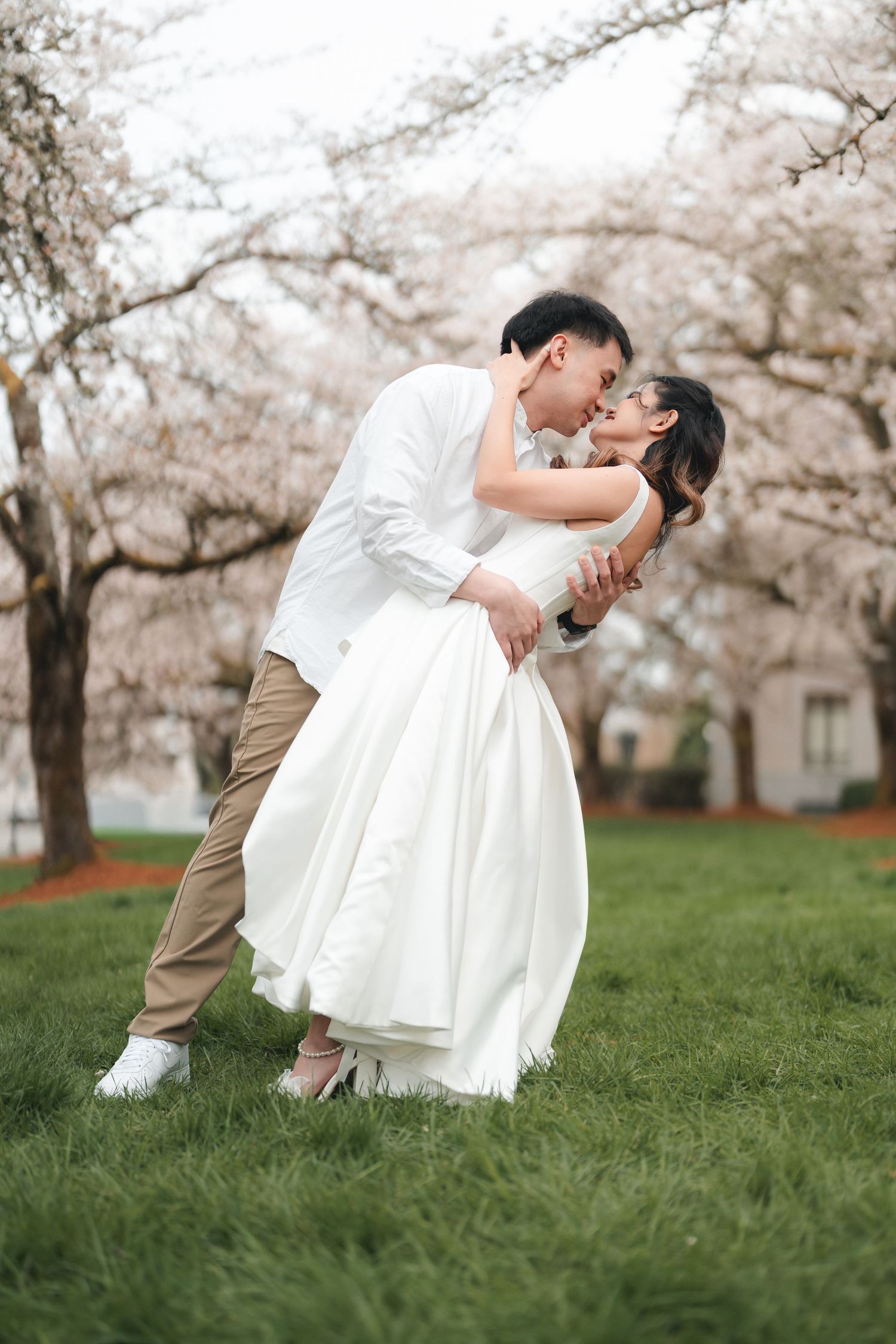 A romantic wedding couple embraces under blooming cherry blossom trees in a spring garden setting.