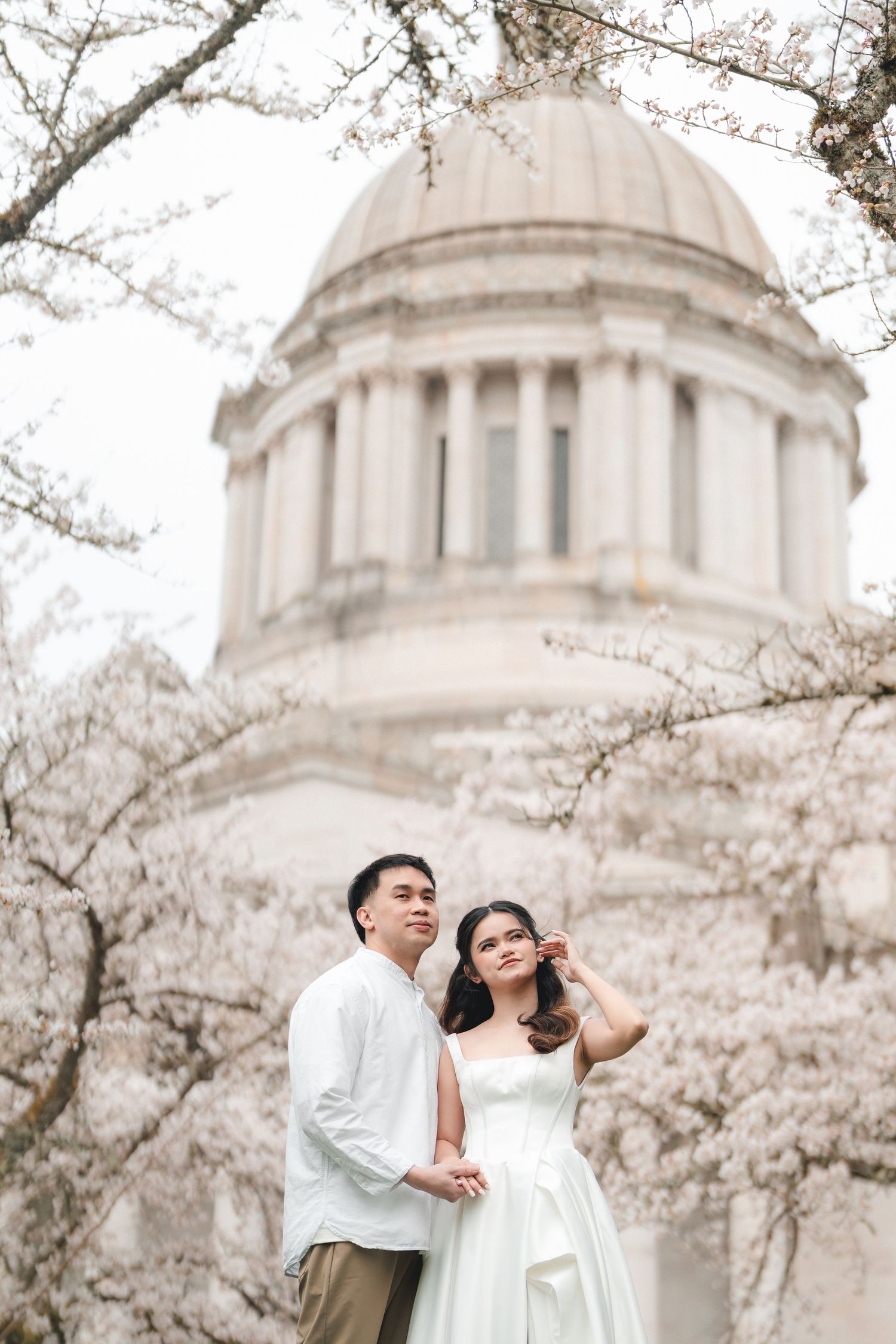 A couple poses romantically among blooming cherry blossoms with a domed building in the background.