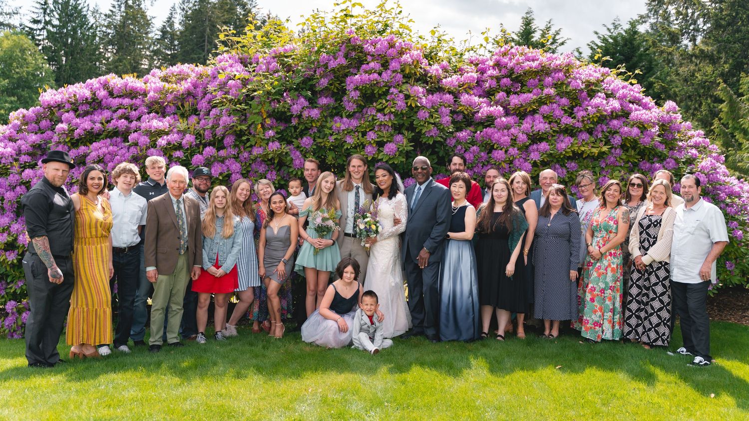 A panoramic view of a group gathering in a garden with blooming pink azalea bushes and evergreen trees in the background.