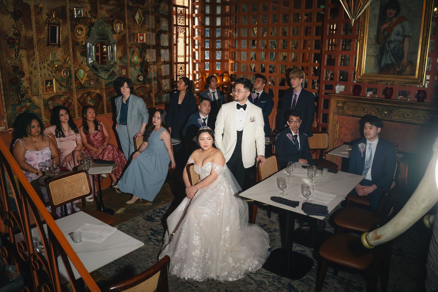 Elegant wedding party poses in ornate restaurant with bride in white gown and groom in tuxedo surrounded by guests.