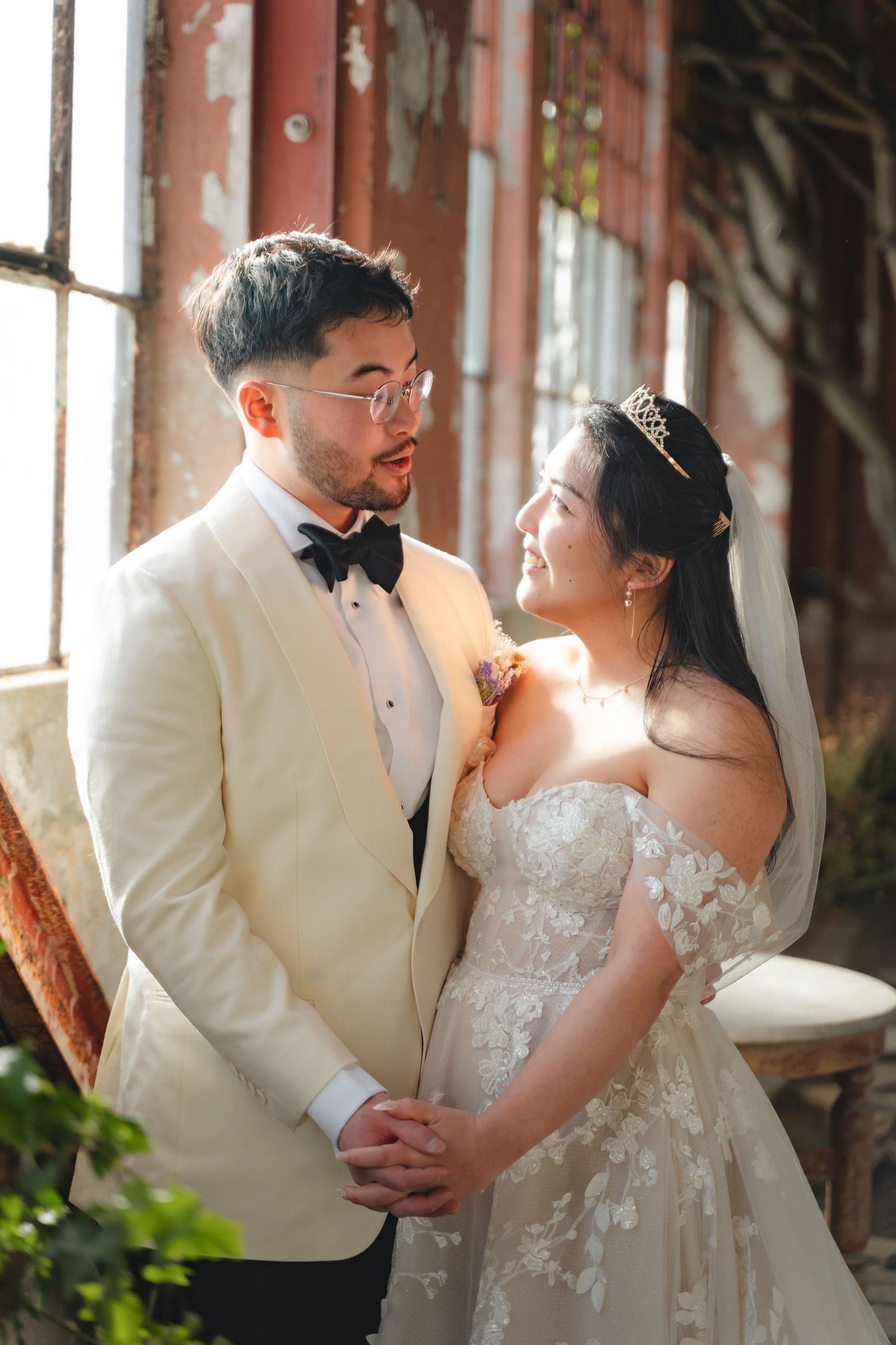 A bride and groom in elegant wedding attire share a tender moment, holding hands by a sunlit window.