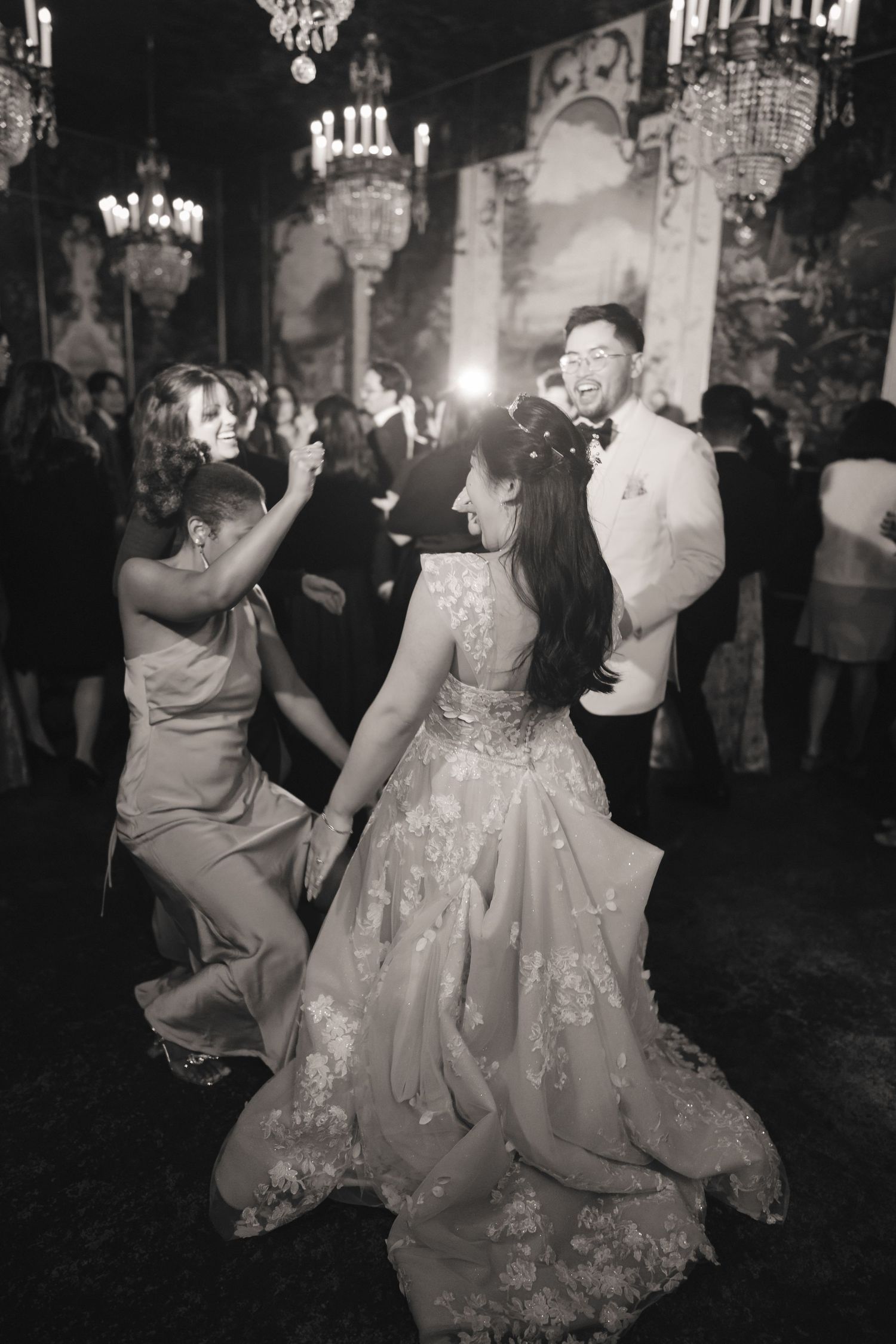 Black and white photo of bride dancing joyfully with guests at elegant wedding reception with crystal chandeliers.