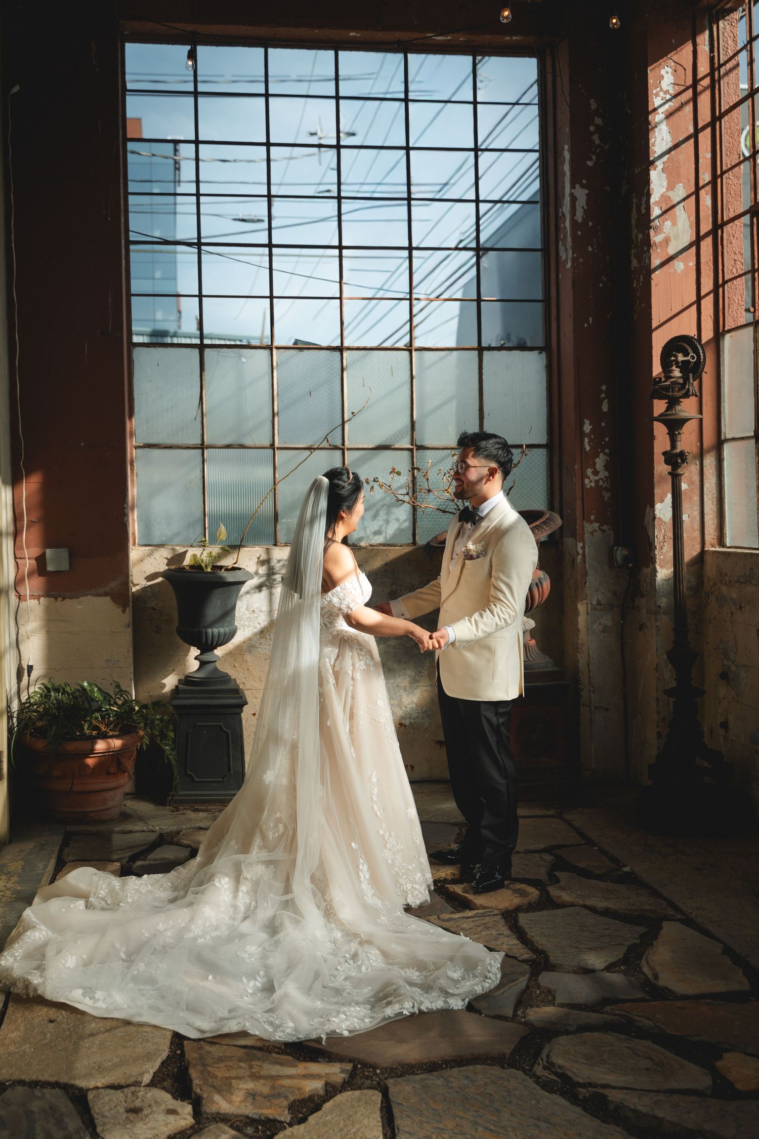 Bride and groom hold hands in a rustic venue with large industrial windows, bride wearing a flowing lace gown with veil.