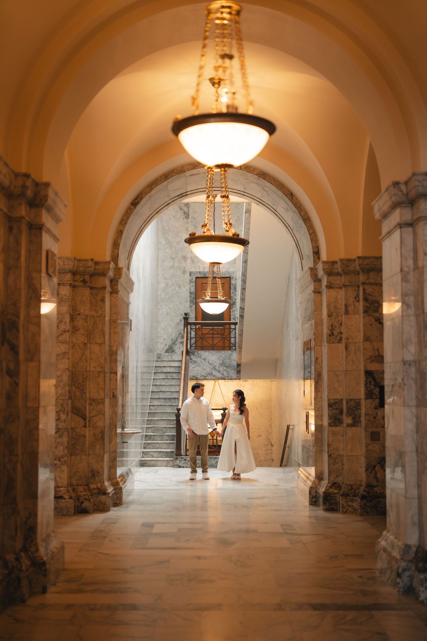 Elegant arched hallway with marble columns, ornate light fixtures, and stone flooring in a historic building interior.