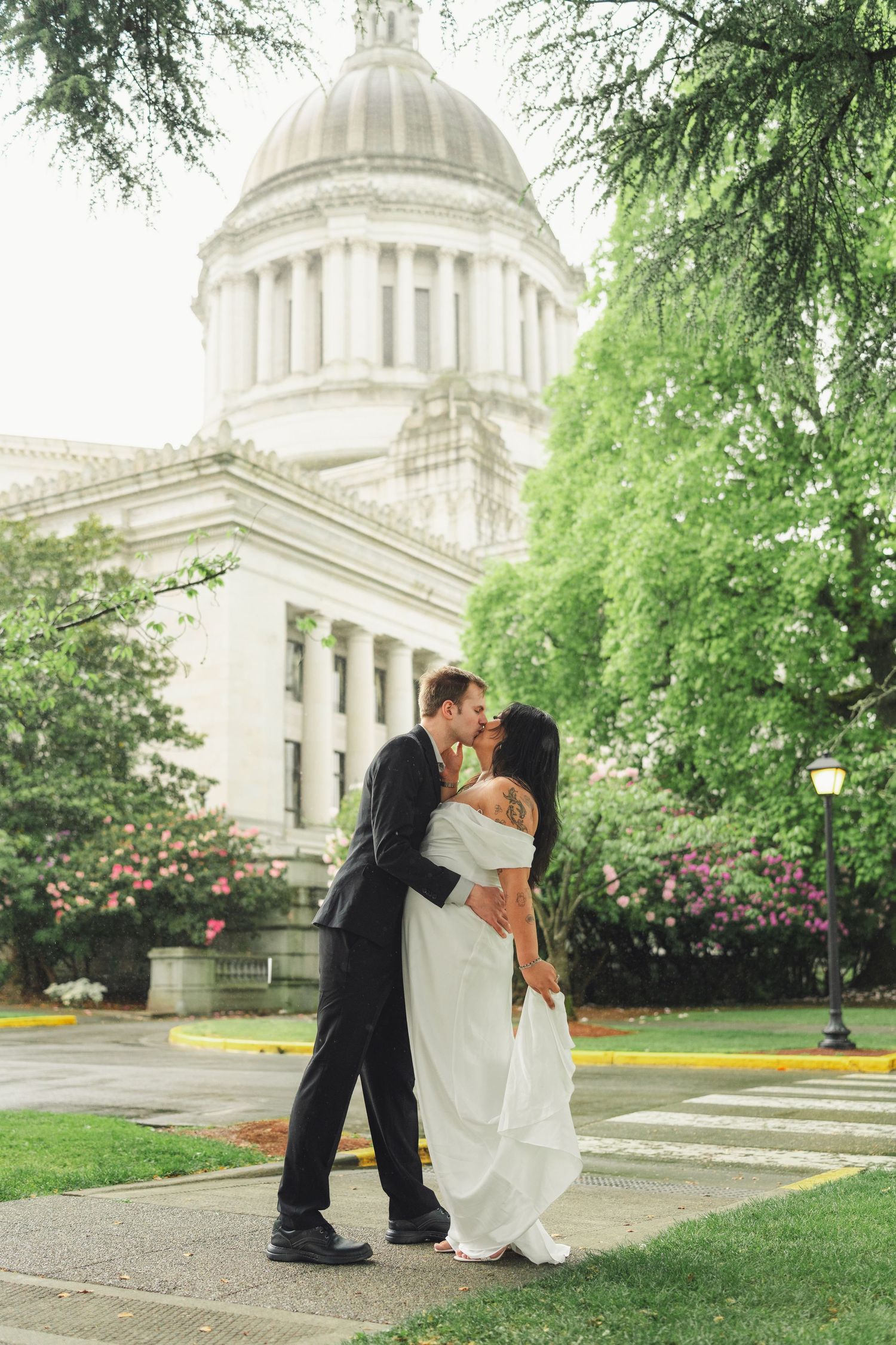 A couple shares a romantic moment in front of a grand domed government building with blooming rose bushes and green trees.