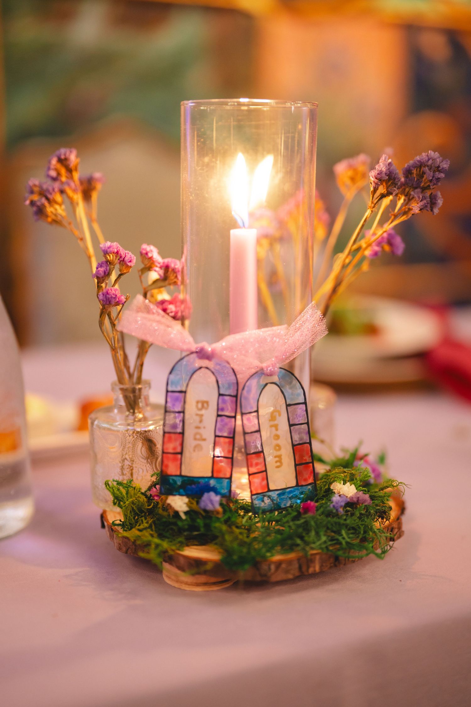 Elegant wedding centerpiece with lit pink candle, dried flowers, and decorative stained glass arch on a mossy wood slice.