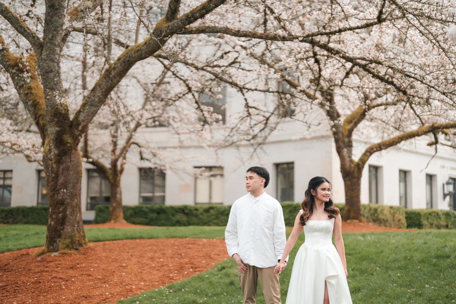 A couple in white outfits strolls through a garden with blooming spring trees and historic buildings in the background.