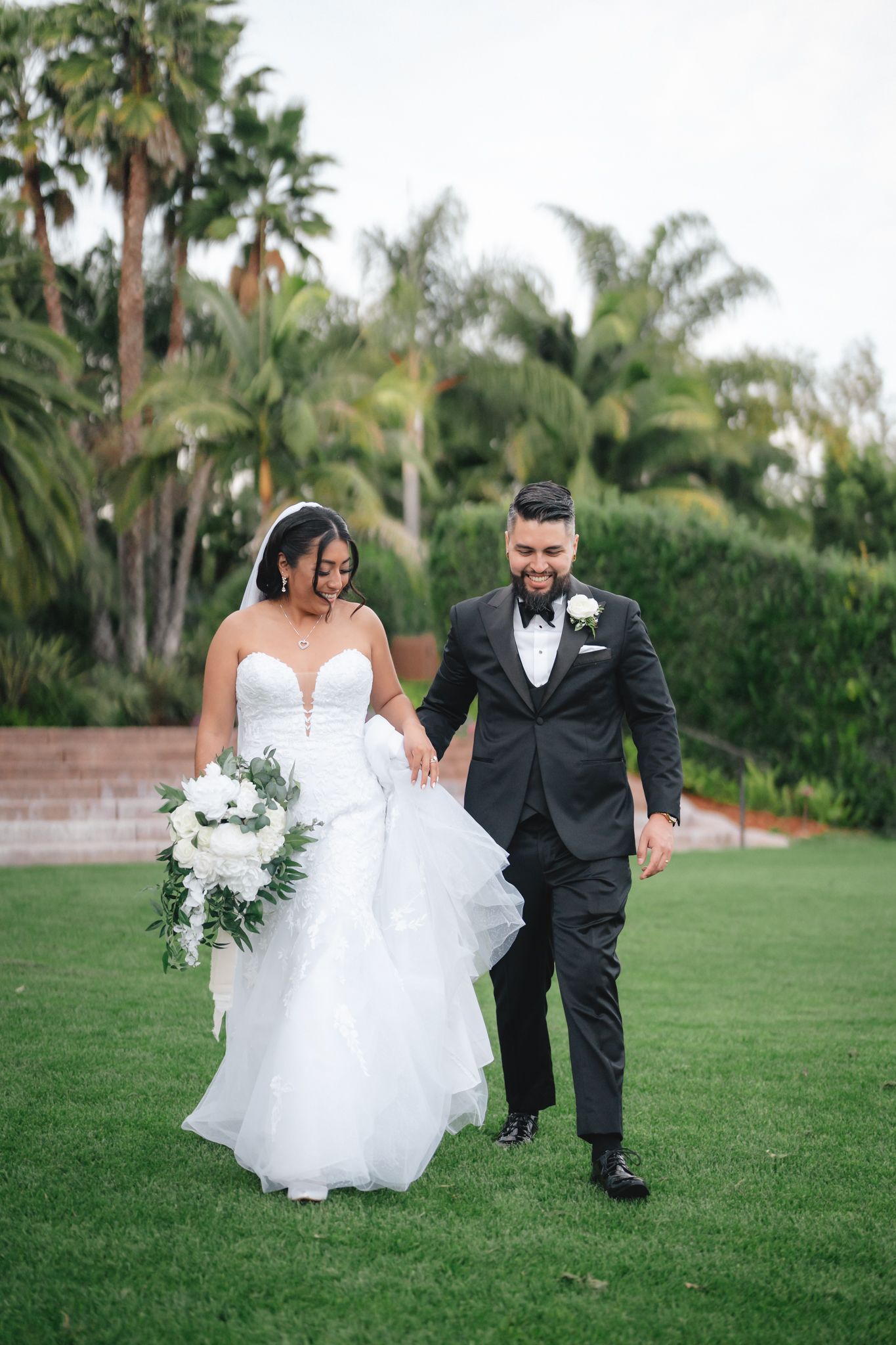 Newlywed couple playfully runs across a lush green lawn with palm trees in the background.