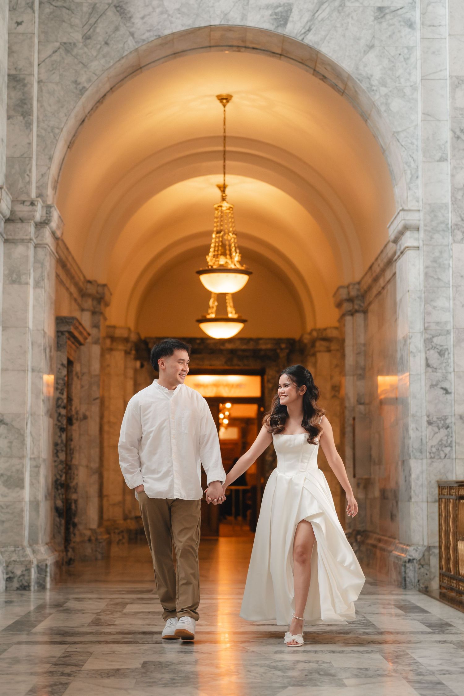 A couple walks hand in hand through an elegant marble hallway with arched ceilings and ornate chandeliers.
