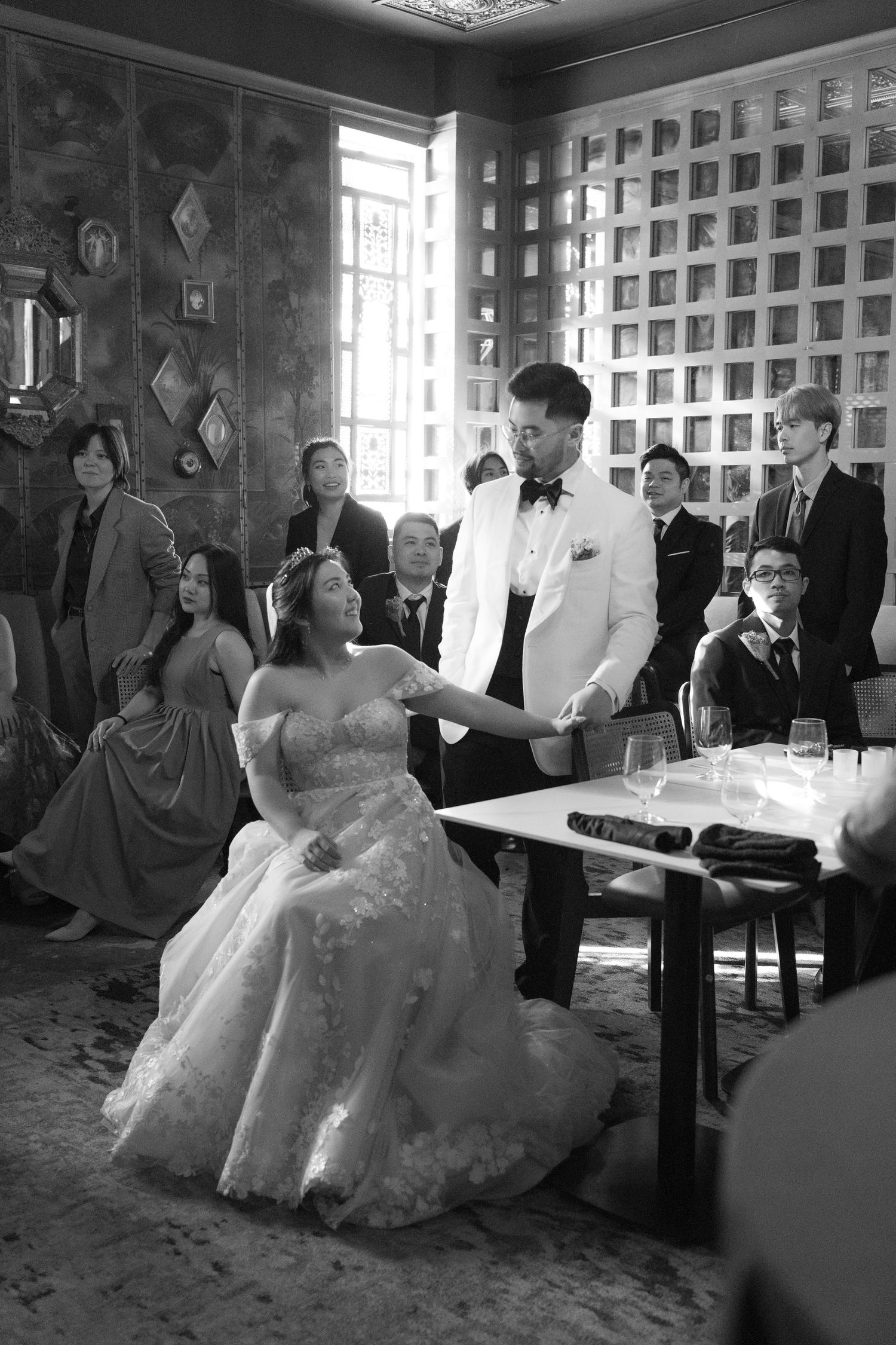 Black and white wedding photo of bride and groom holding hands at reception table with guests seated behind them.