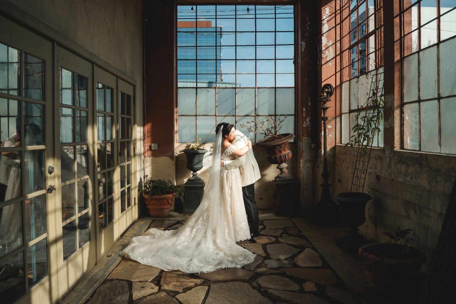 Bride in elegant white gown and cape standing in sunlit rustic venue with stone floors and large windows.