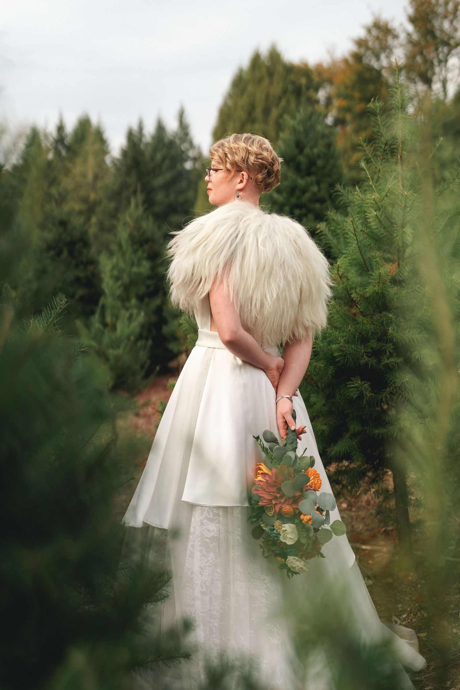 Two people in white wedding attire embrace in a romantic outdoor garden setting surrounded by evergreen trees.