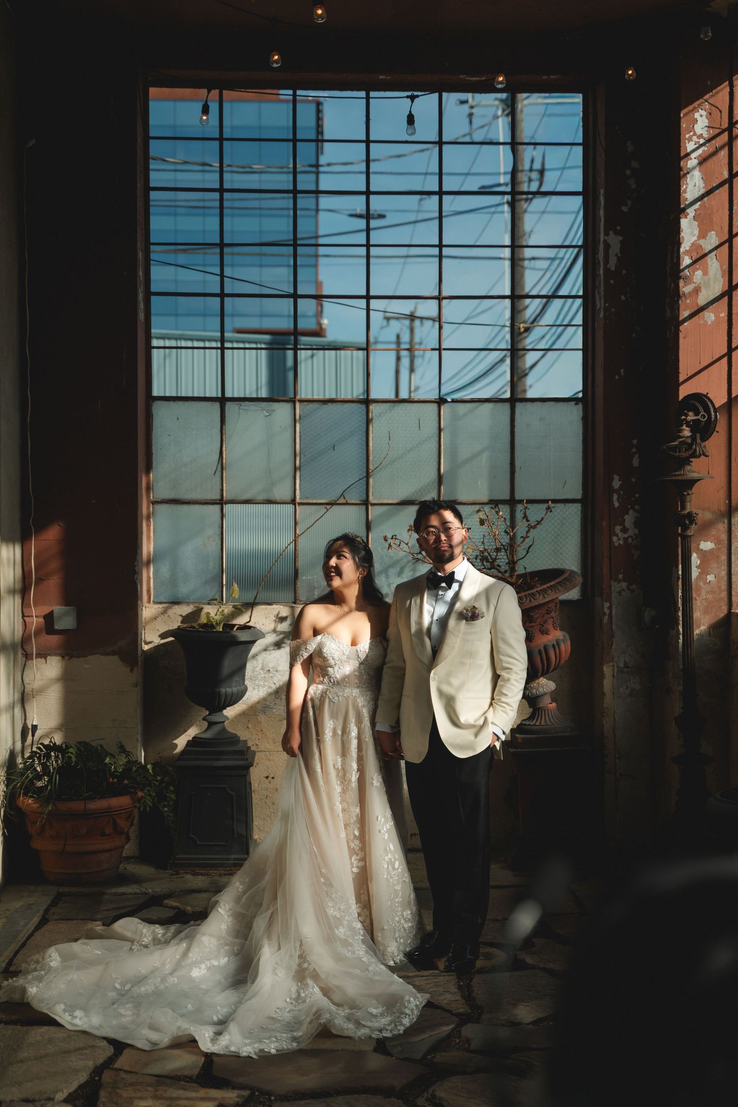 Bride and groom pose in elegant wedding attire before a large industrial window with warm sunlight.