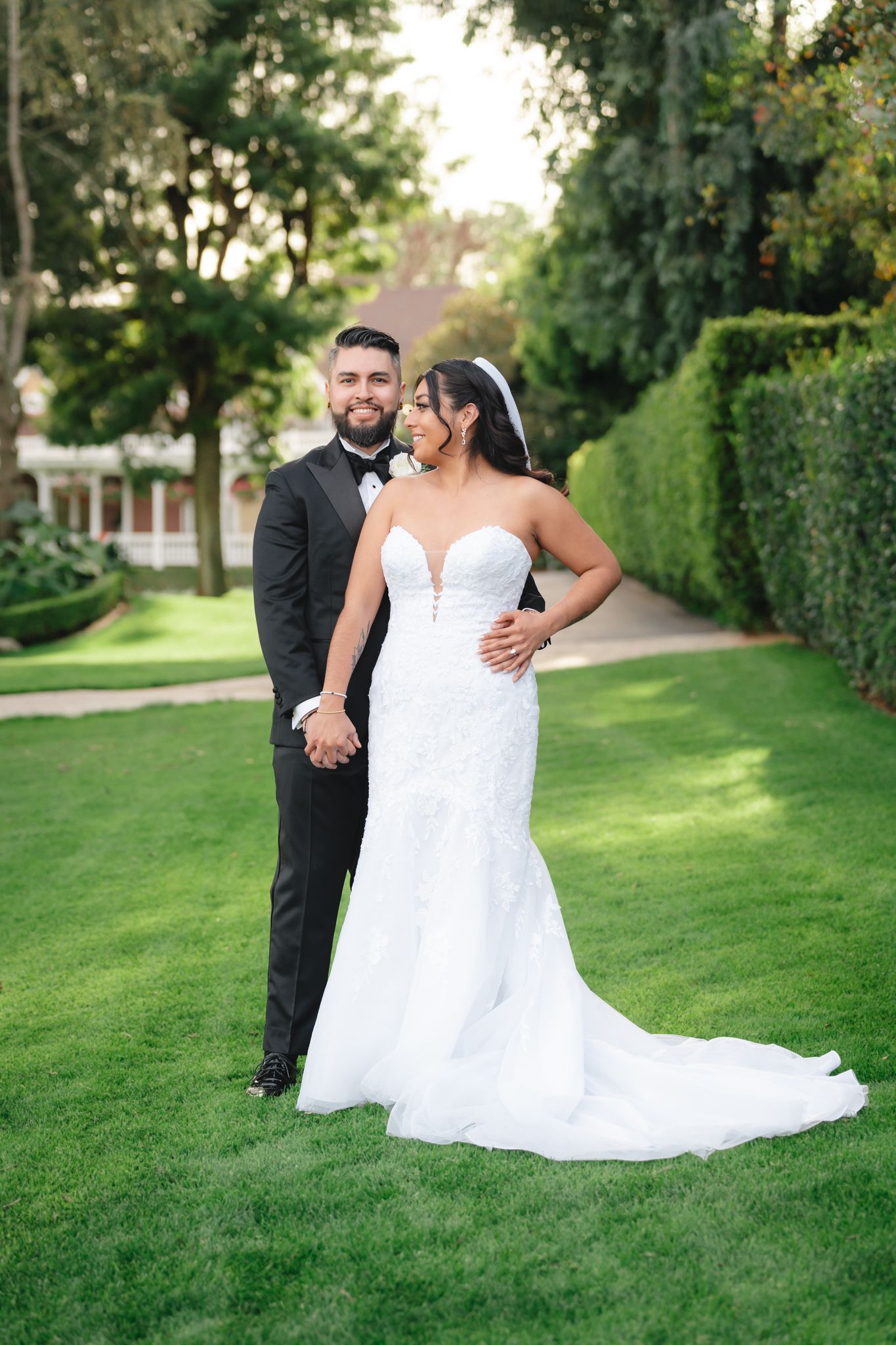Wedding couple poses together on a lush green lawn with elegant landscaping in the background.