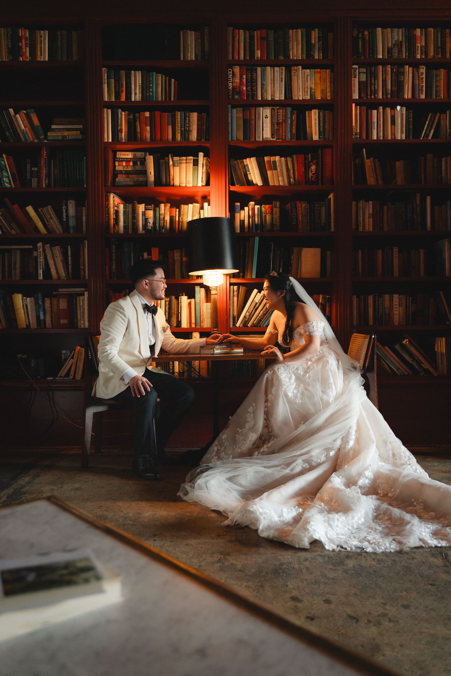 Bride and groom sit together in a warmly lit library surrounded by bookshelves, sharing an intimate moment on their wedding day.