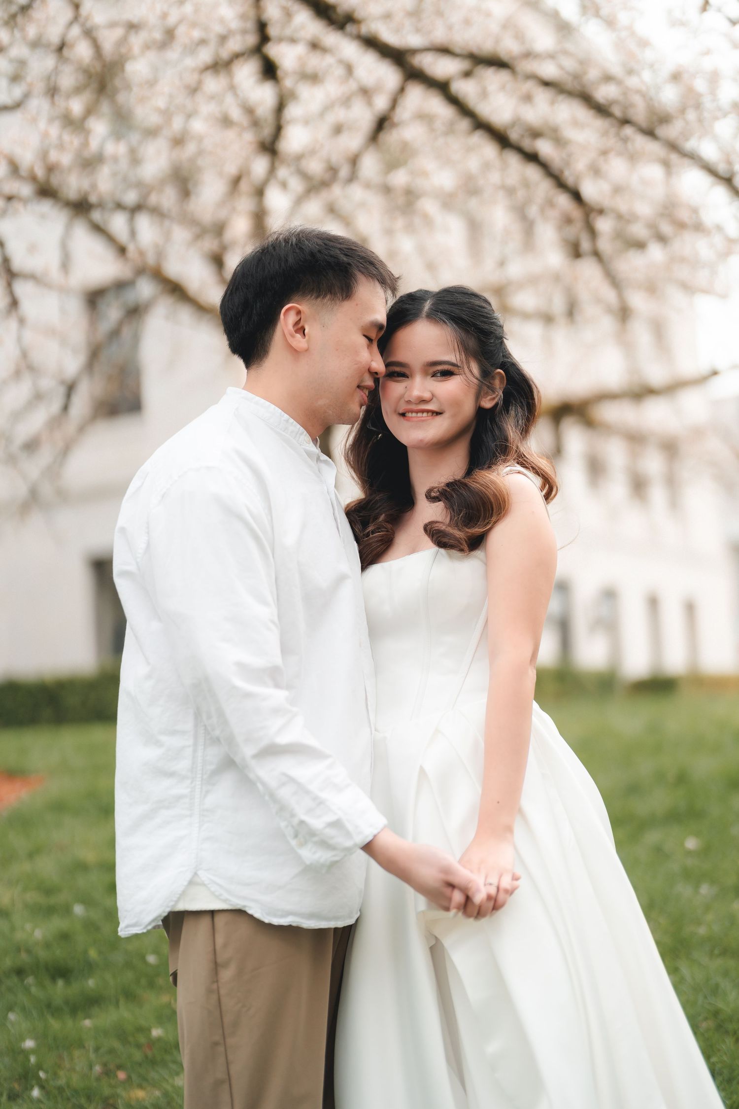 A romantic couple in white attire share an intimate moment under blooming cherry blossom trees in a garden setting.