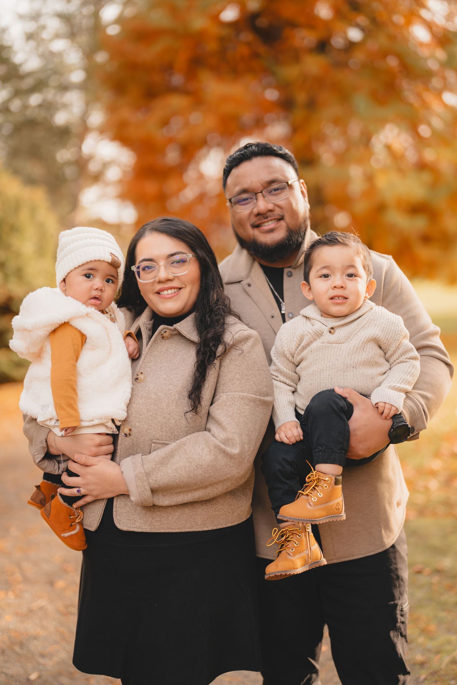 A family wearing coordinating beige sweaters poses together against a backdrop of autumn foliage.