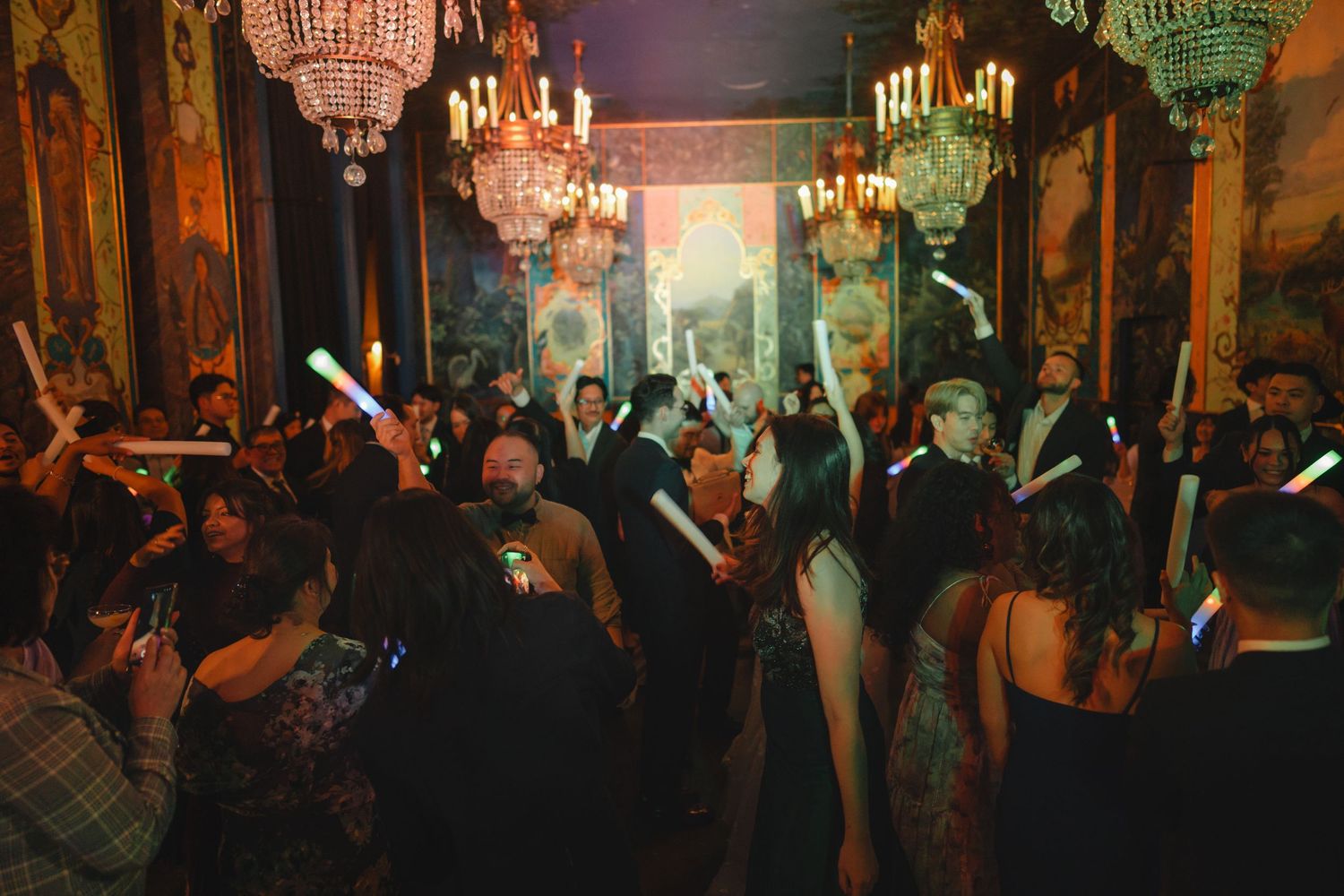 Elegant ballroom party with guests holding glow sticks under crystal chandeliers in a ornate golden venue.