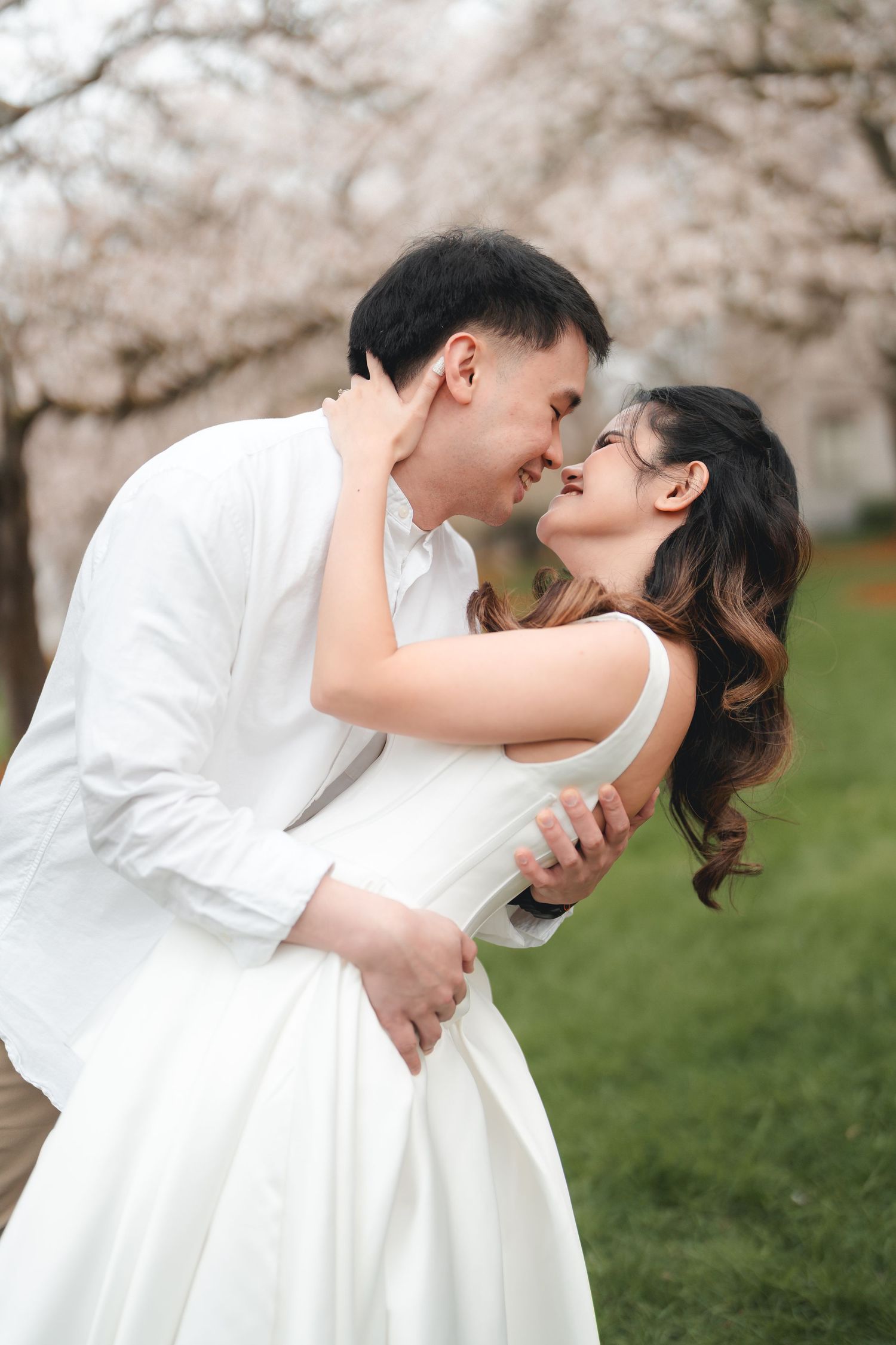 A romantic couple in white attire share an intimate moment under blooming cherry blossom trees in a garden setting.