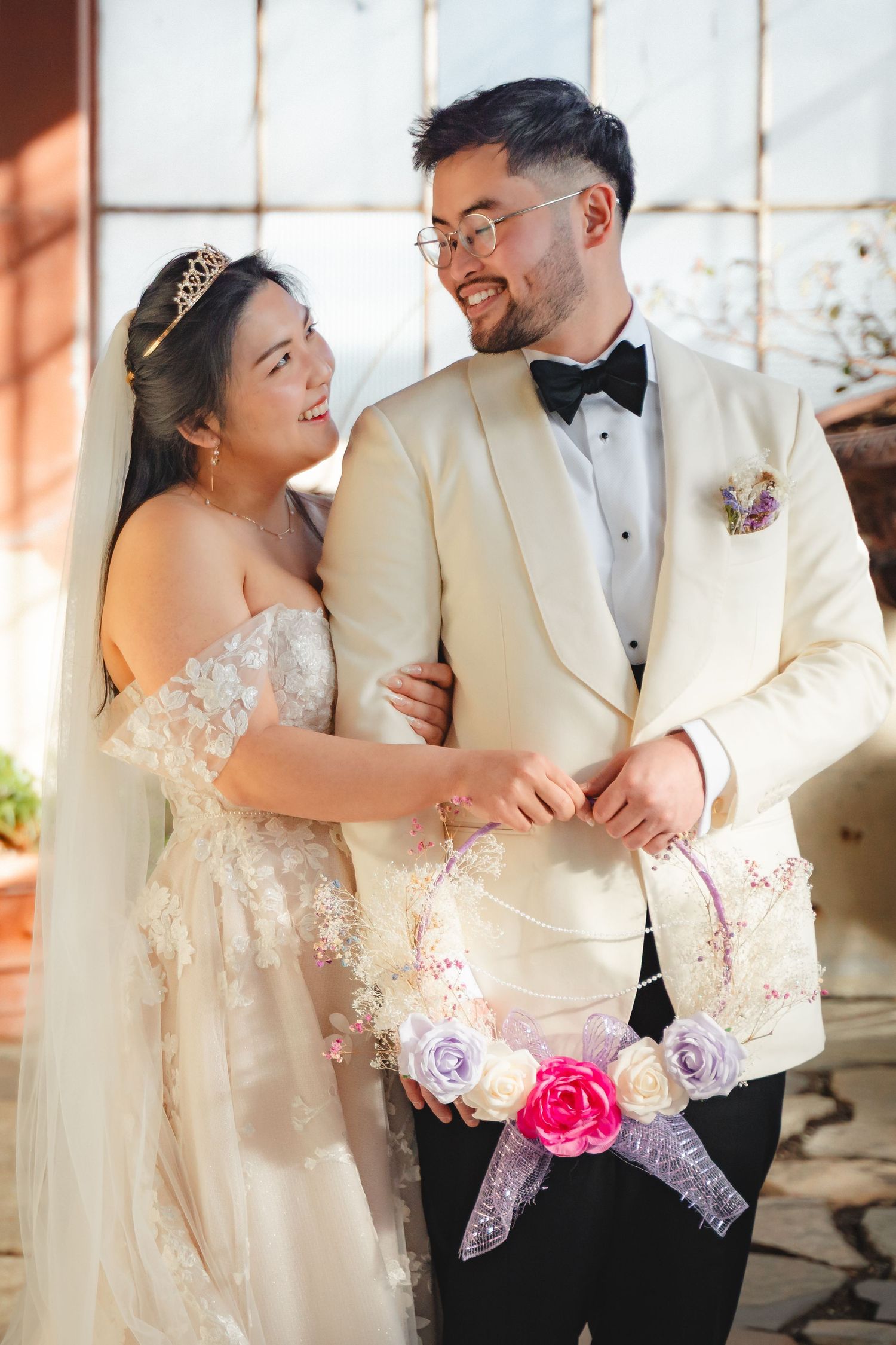 Bride and groom smiling at each other, holding floral wreath, bride in white gown, groom in cream tuxedo with bow tie.