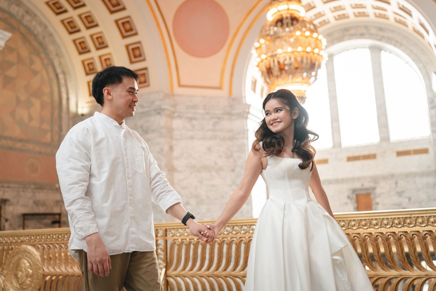 Couple in elegant white attire share romantic moments in an ornate historic building with grand architecture and chandeliers.