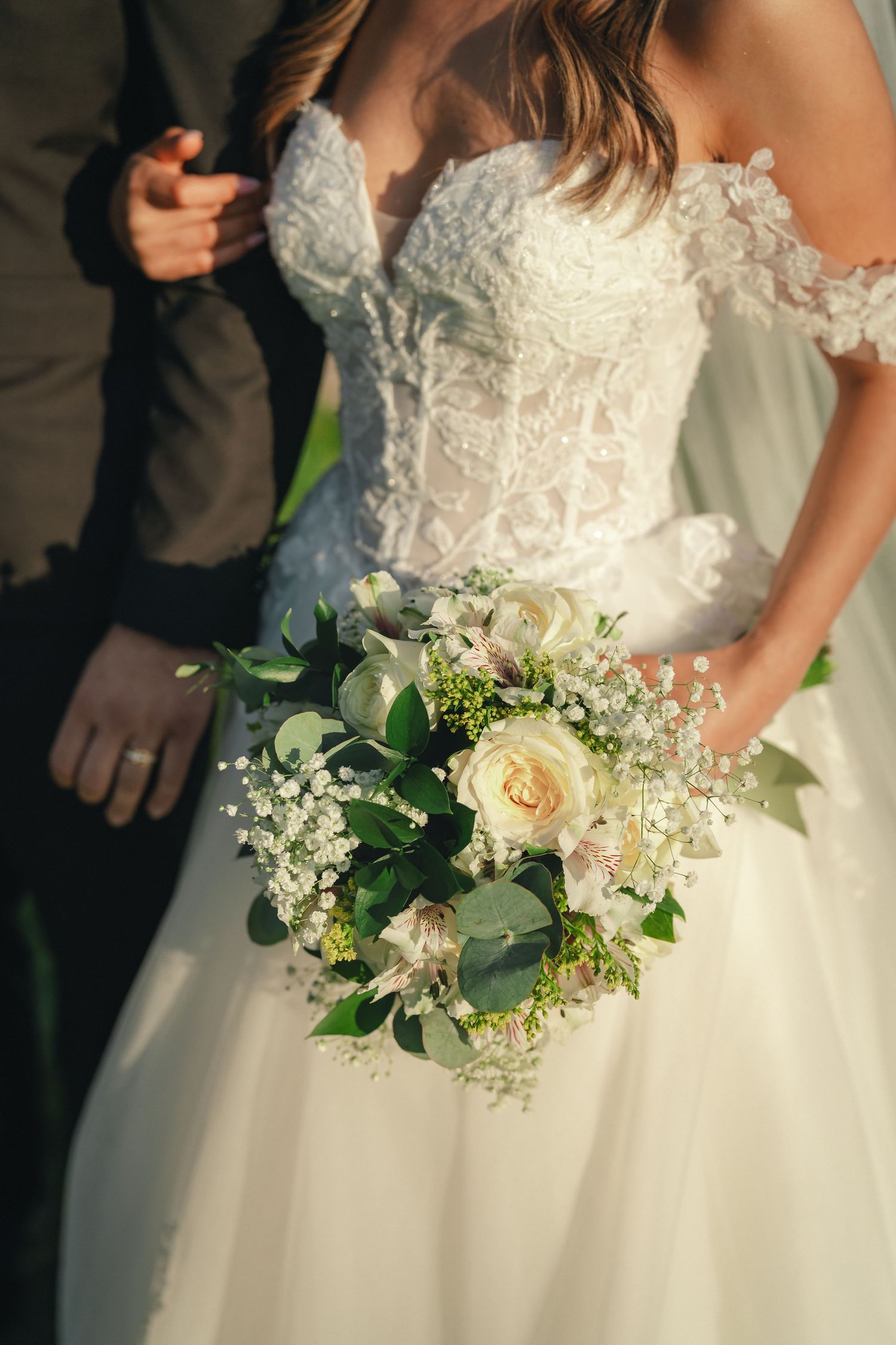 A white wedding bouquet featuring cream roses, delicate baby's breath, and green eucalyptus leaves against a lace gown.