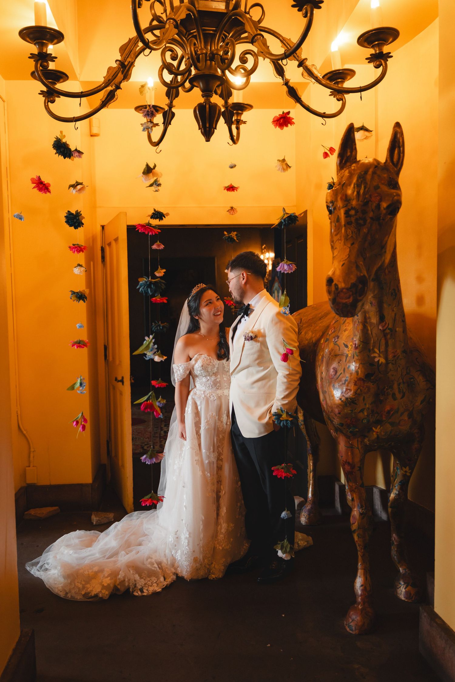Bride and groom stand together in a warmly lit hallway with a chandelier, floral garlands, and a decorative horse statue.