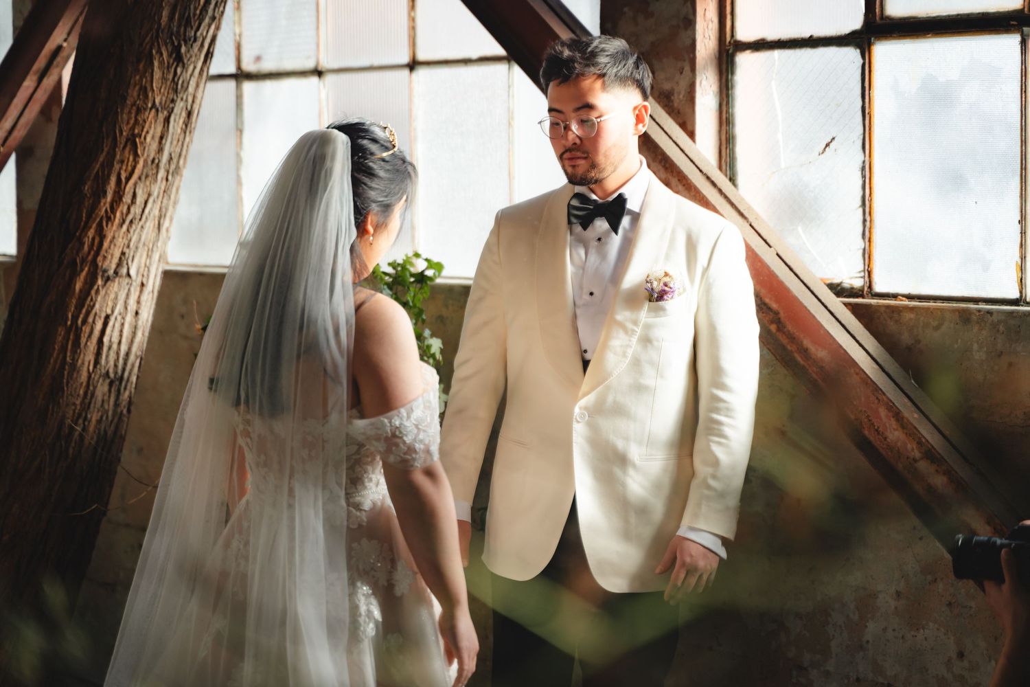 Bride and groom share an intimate moment in a rustic venue, with bride in veil and groom in white tuxedo.