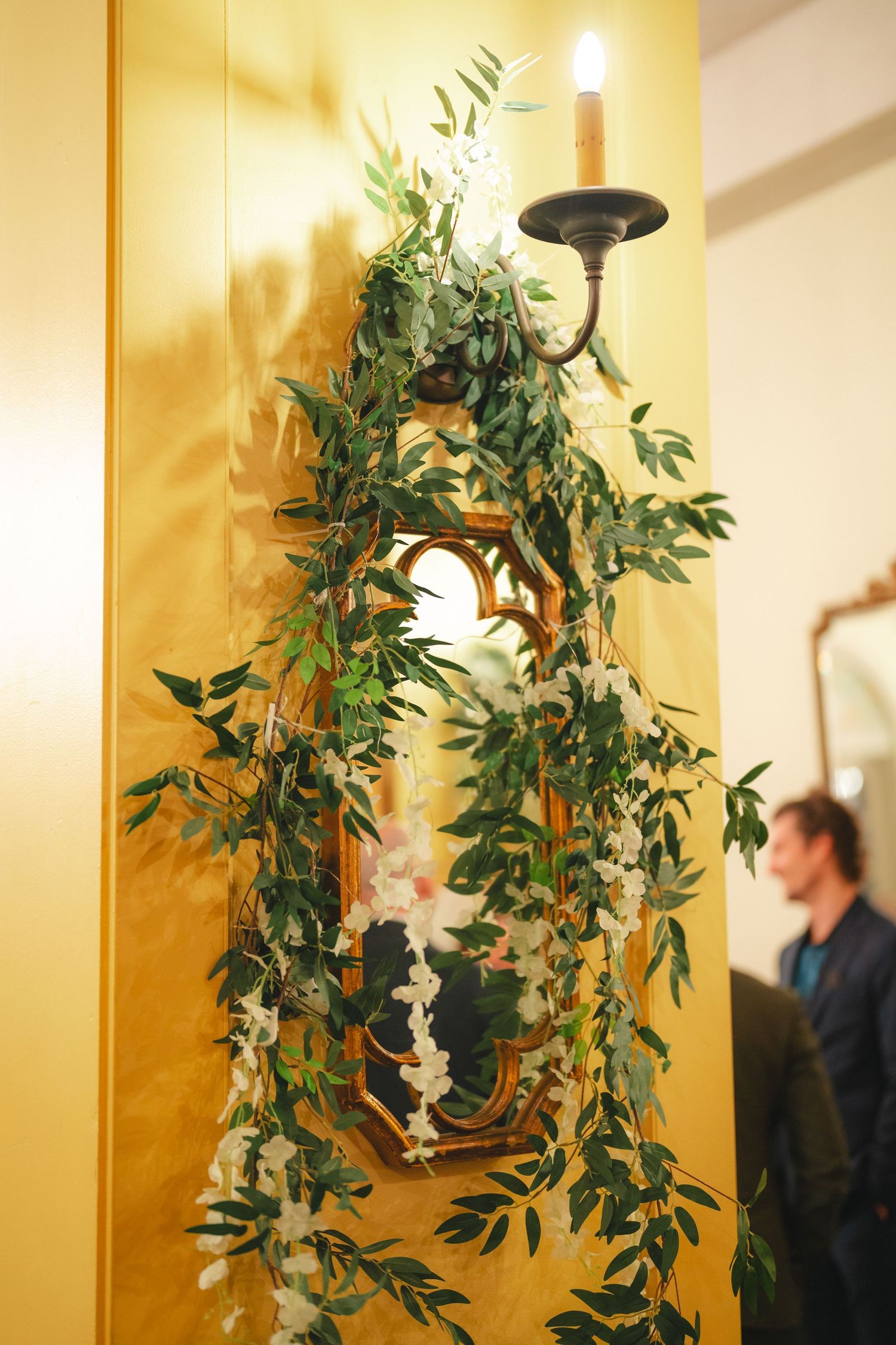 Gold-framed mirror adorned with lush green garlands and white flowers, topped with a black wall sconce candle holder.