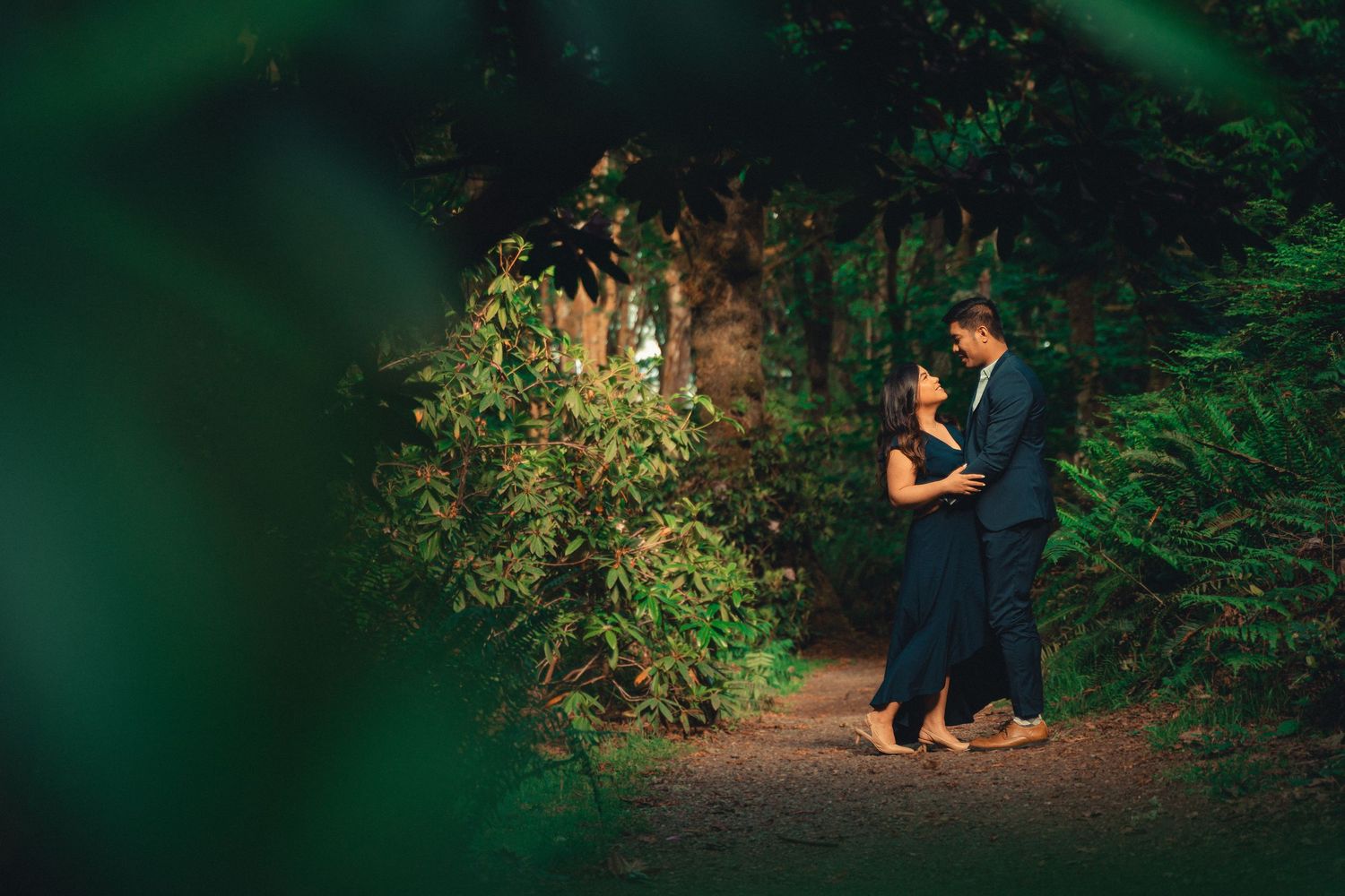 A romantic couple shares a tender moment together on a wooded path illuminated by soft natural light.