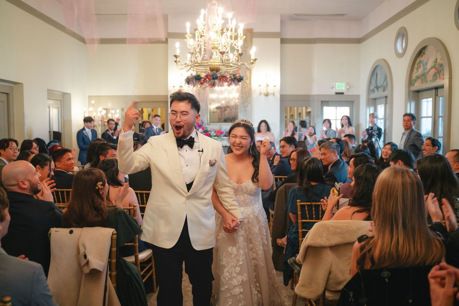 Newlywed couple walks down the aisle at elegant indoor wedding reception with chandelier and applauding guests.