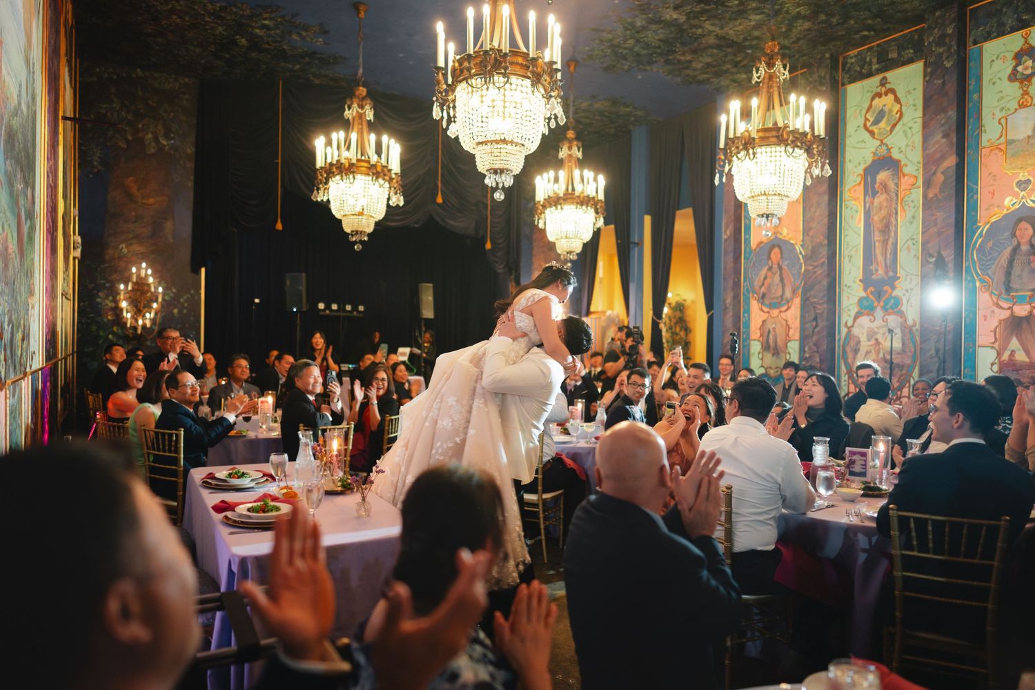 Couple sharing a romantic kiss at an elegant wedding reception in a grand ballroom with chandeliers and guests applauding.